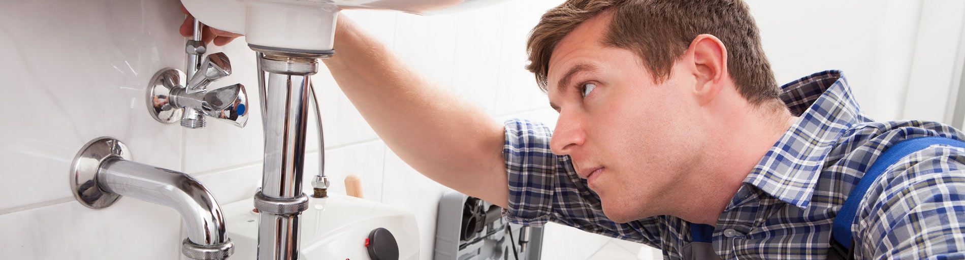 Fontanero arreglando tuberías debajo de un fregadero. Lleva una camisa azul a cuadros y se concentra intensamente.