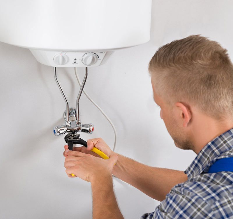 Una persona usa alicates para trabajar en accesorios de plomería debajo de un calentador de agua blanco montado en una pared.