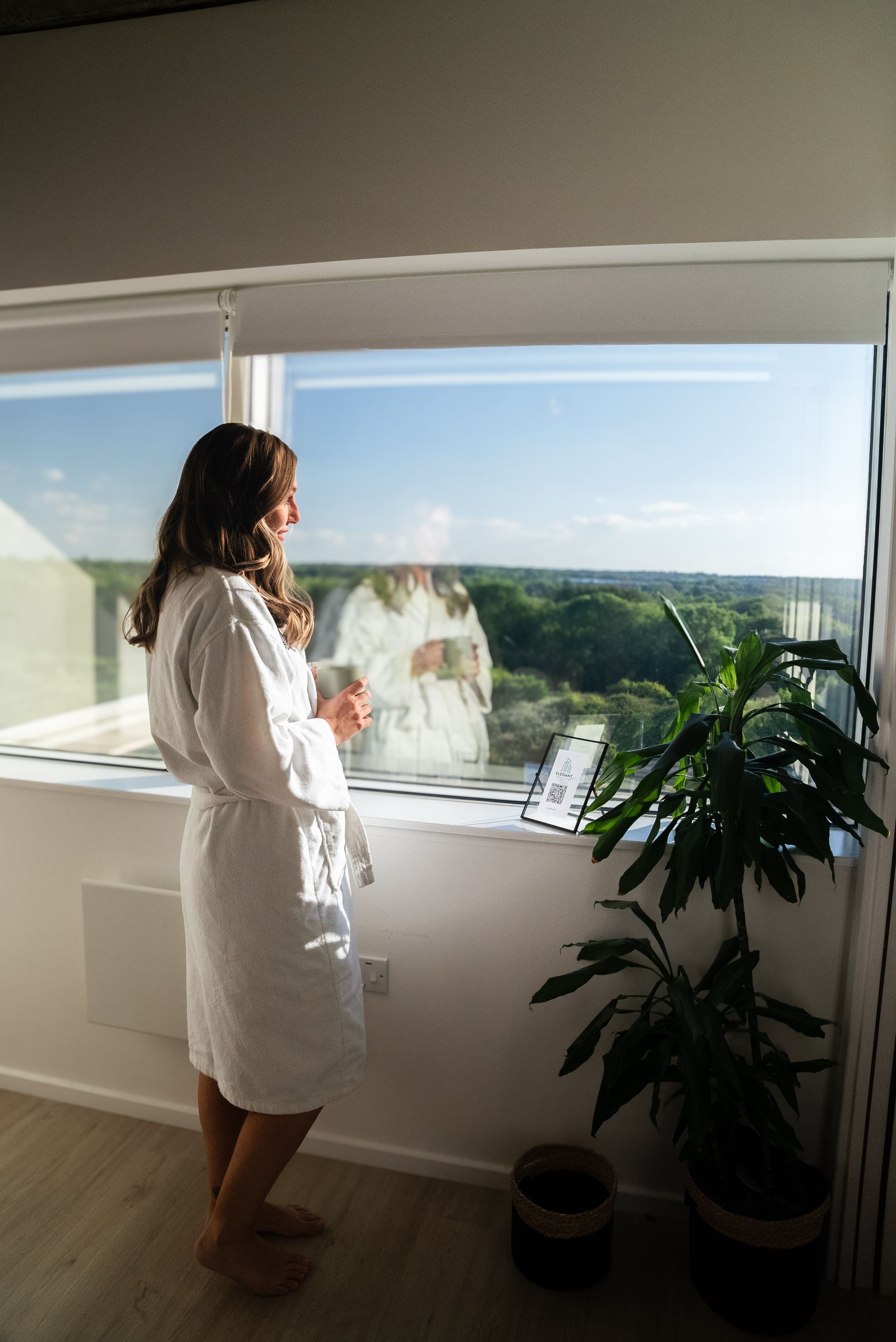 Woman in robe looking out window, holding mug, standing near plant. Bright sunlight.