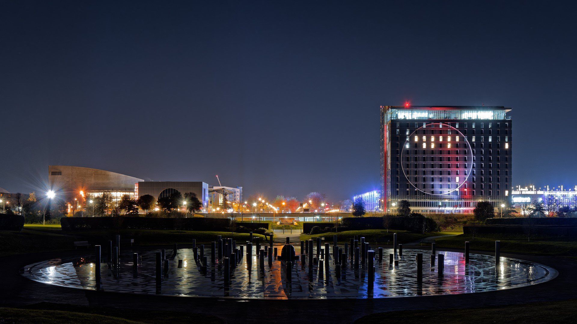 Night view of a cityscape with illuminated buildings reflected in a dark water feature.