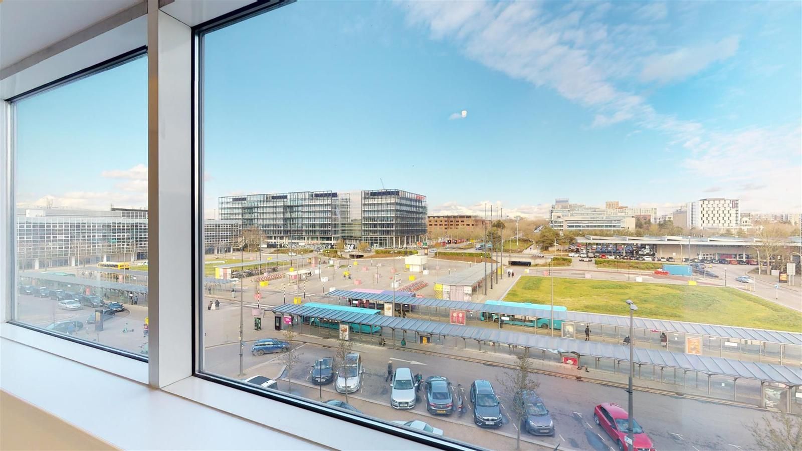 View from a window overlooking a city square with buildings, pedestrians, and cars on a sunny day.