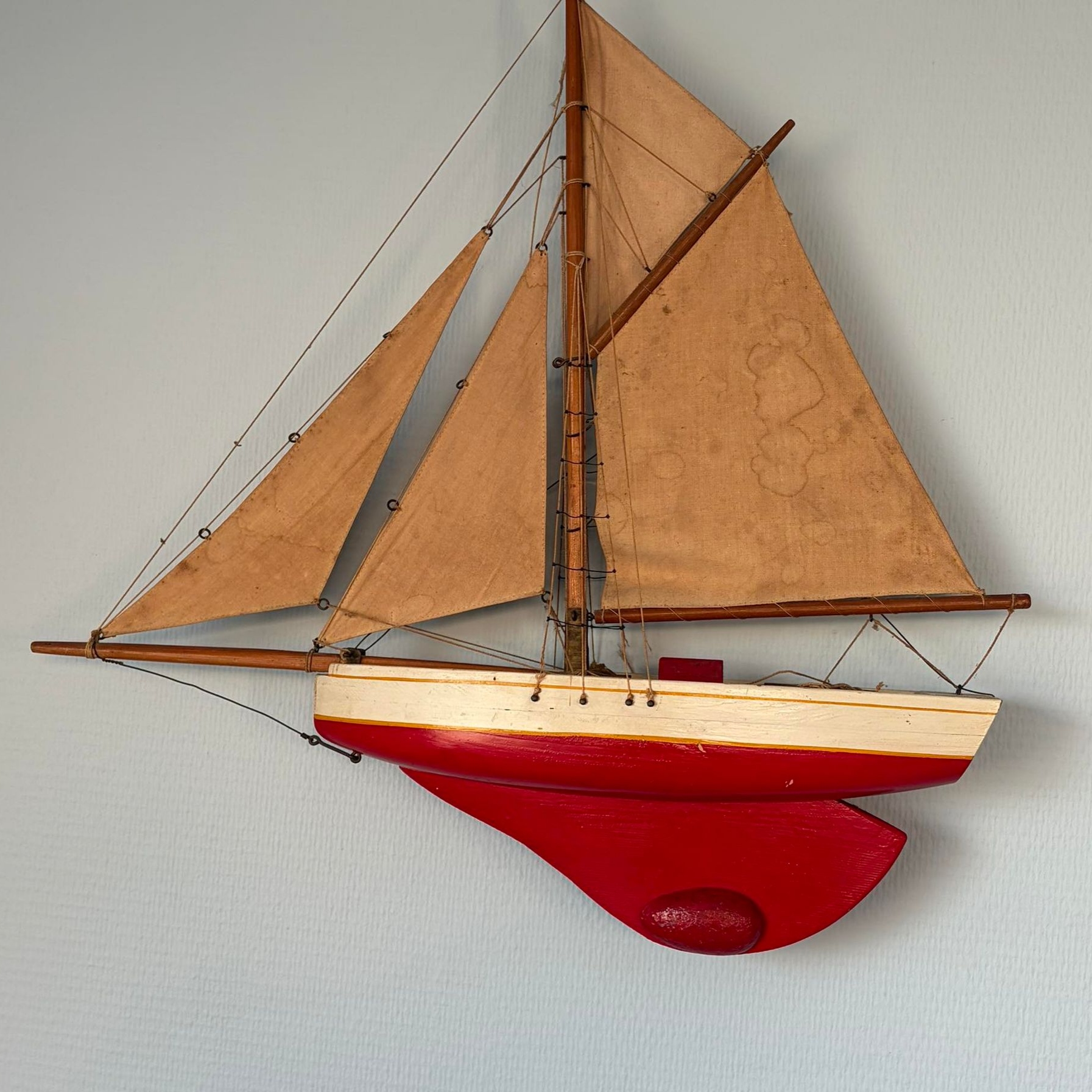 Un petit modèle réduit de voilier en bois, avec des voiles beiges, une coque crème et un socle rouge, fixé au mur.