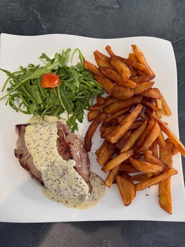 Pièce de viande servie dans une assiette blanche, nappée d'une sauce crémeuse, accompagné de salade de roquette et de frites croustillantes.