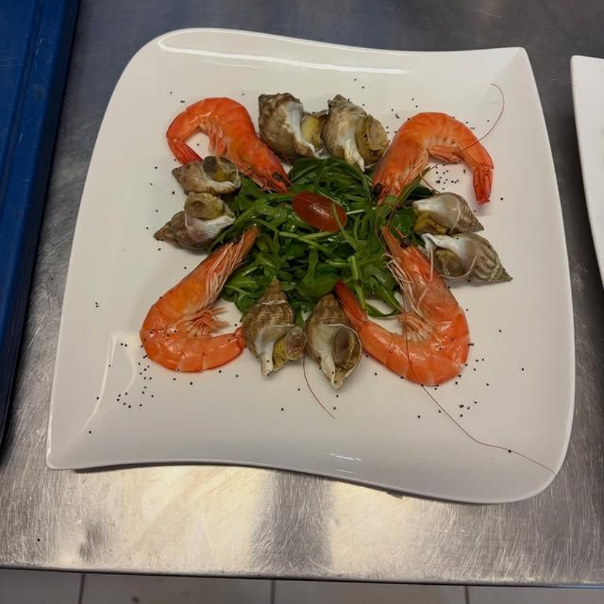 Crevettes et fruits de mer disposés autour de verdure sur une assiette carrée blanche, arrosés de sauce
