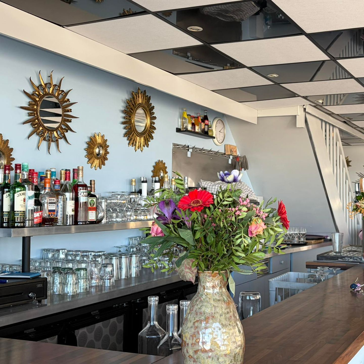 Un bar avec des bouteilles d'alcool, de la verrerie et un vase de fleurs roses sous un décor mural en forme de soleil.
