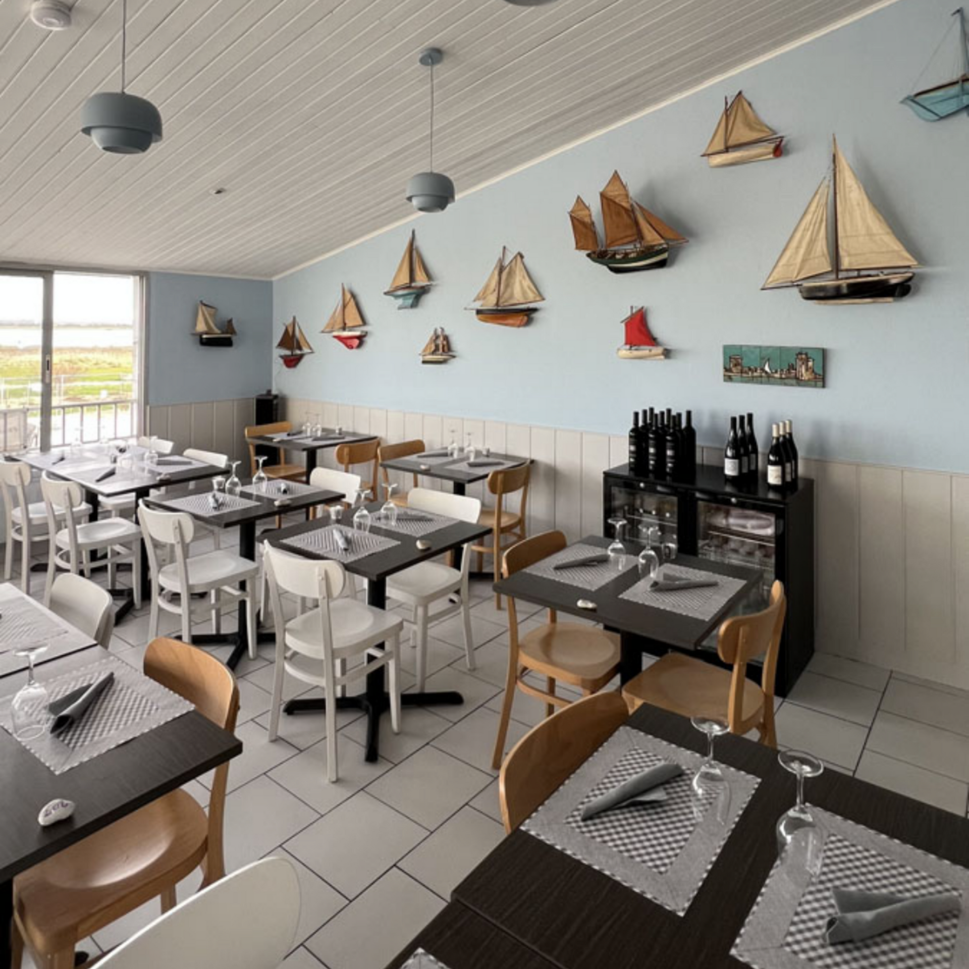 Salle à manger lumineuse d'un restaurant en bord de mer, décorée de voiliers, tables dressées et vue sur l'eau par la fenêtre.