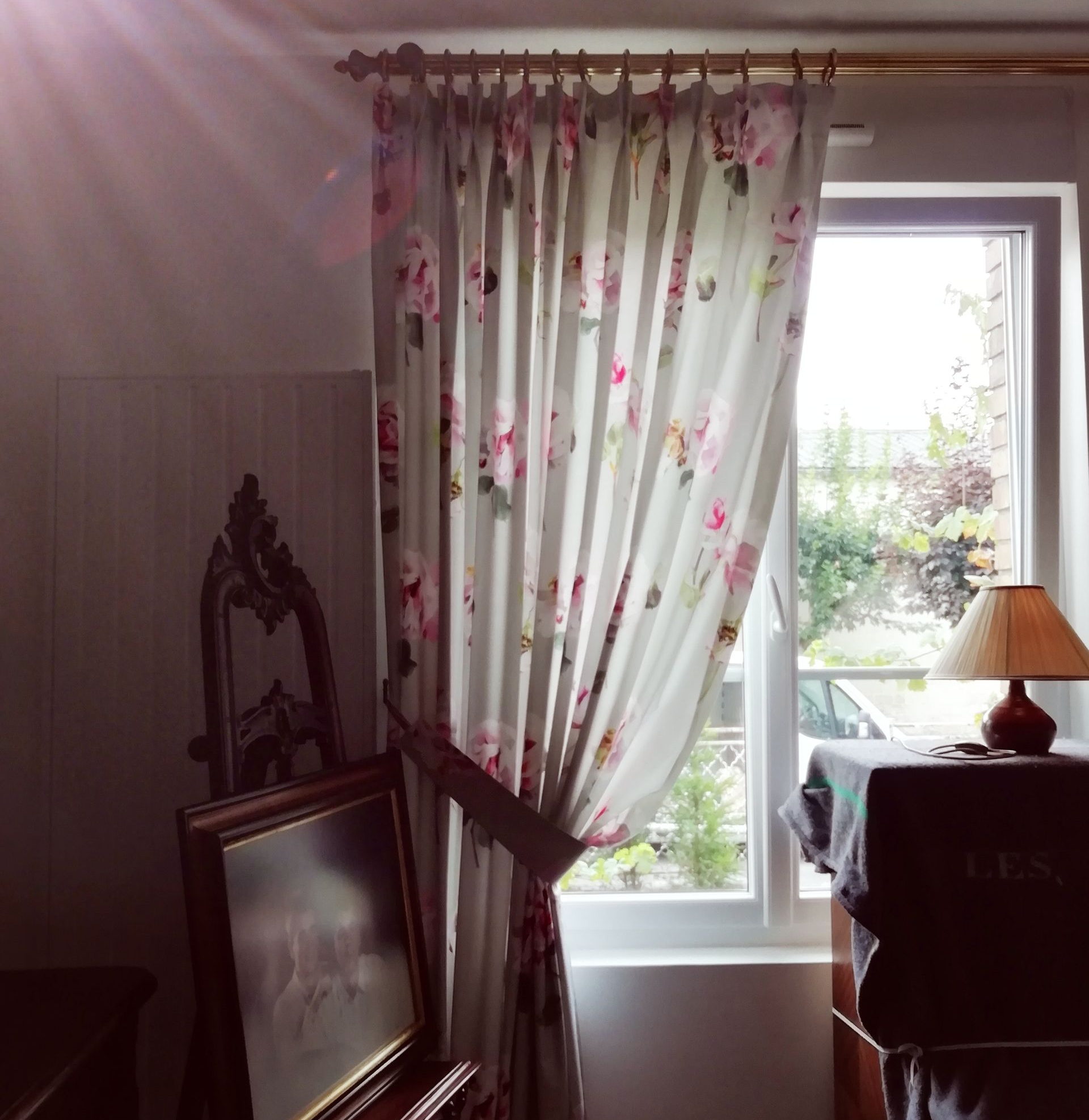 Une fenêtre avec des rideaux à fleurs, un tableau et une lampe dans une pièce baignée de soleil.