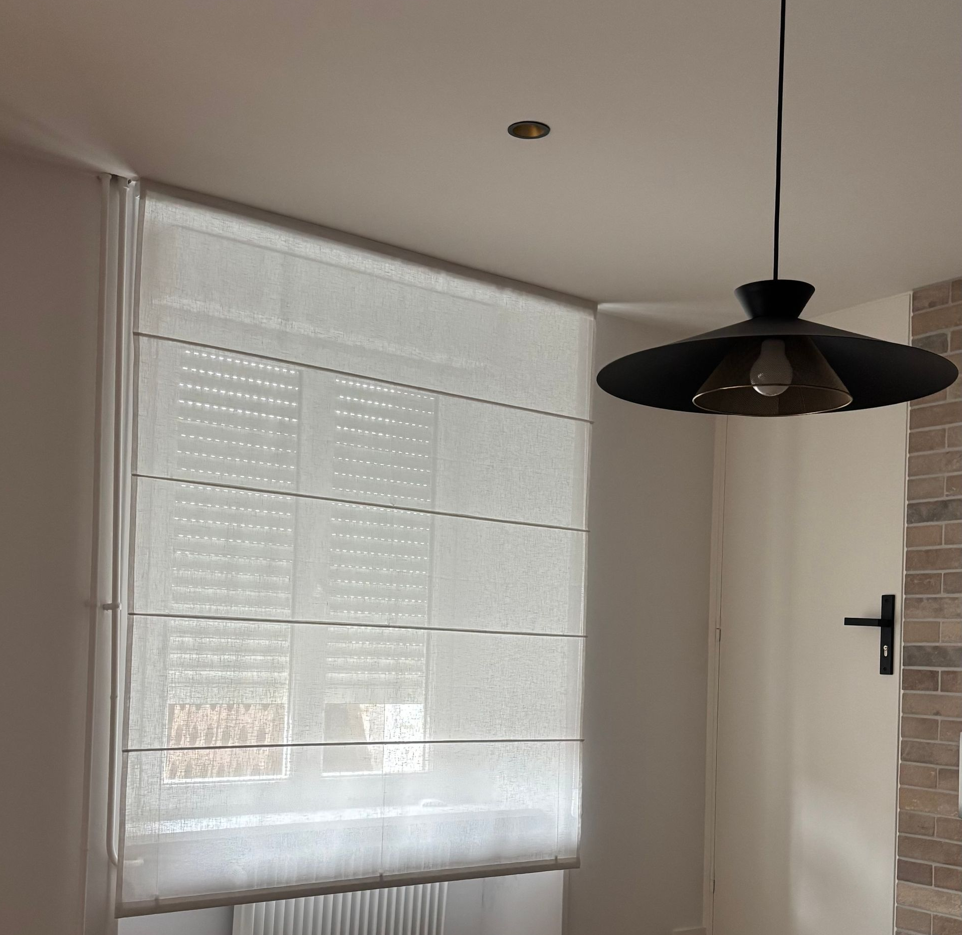 Stores à rayures zébrées grises et blanches dans une fenêtre à cadre blanc donnant sur un paysage urbain.