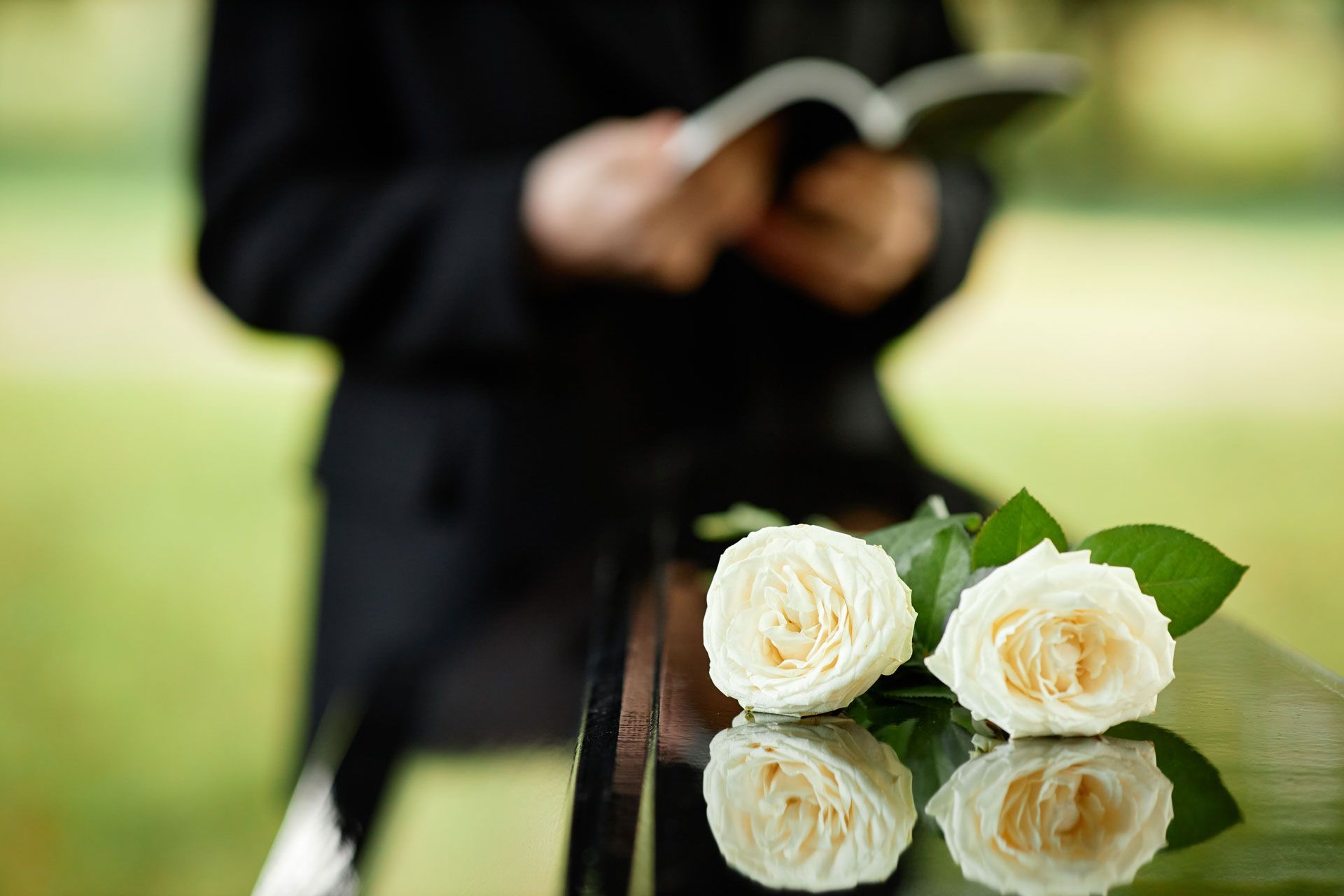 Plan rapproché sur des roses blanches posées sur un cercueil, en fond un homme d'église lisant la Bible