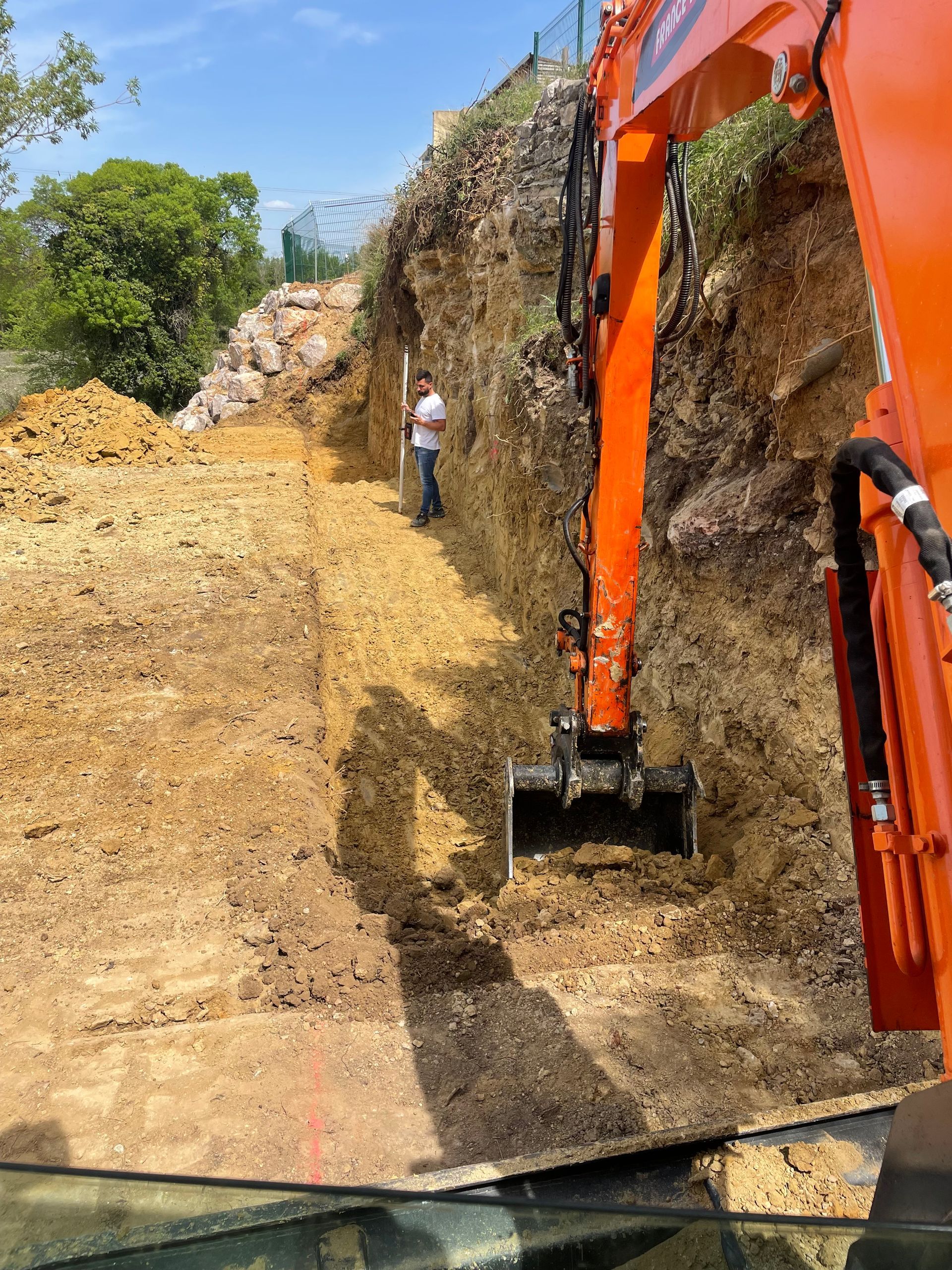 Une pelleteuse orange creuse un talus de terre à côté d'une colline rocheuse.