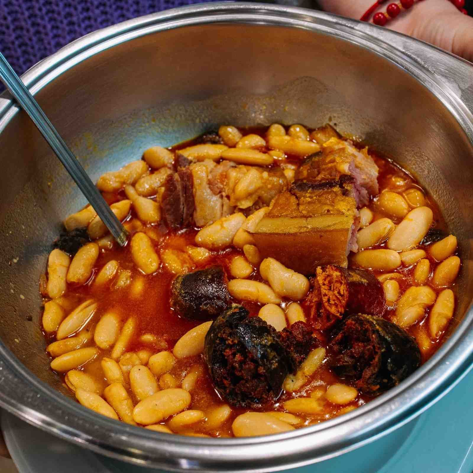 Un tazón de metal con sopa de frijoles, trozos de cerdo y una cuchara sobre una mesa.