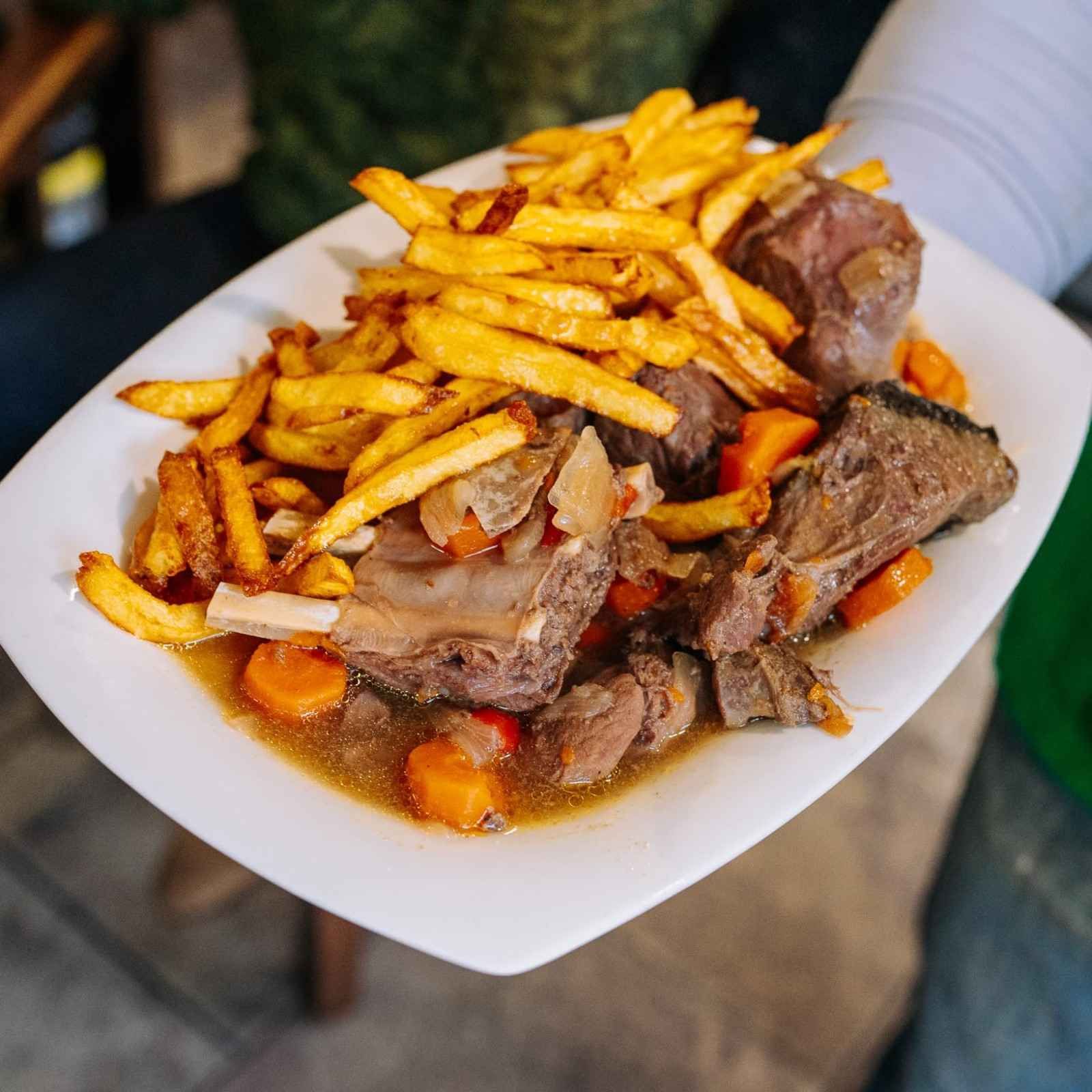 Plato de patatas fritas y carne asada con verduras en un plato blanco, sostenido en las manos de alguien.