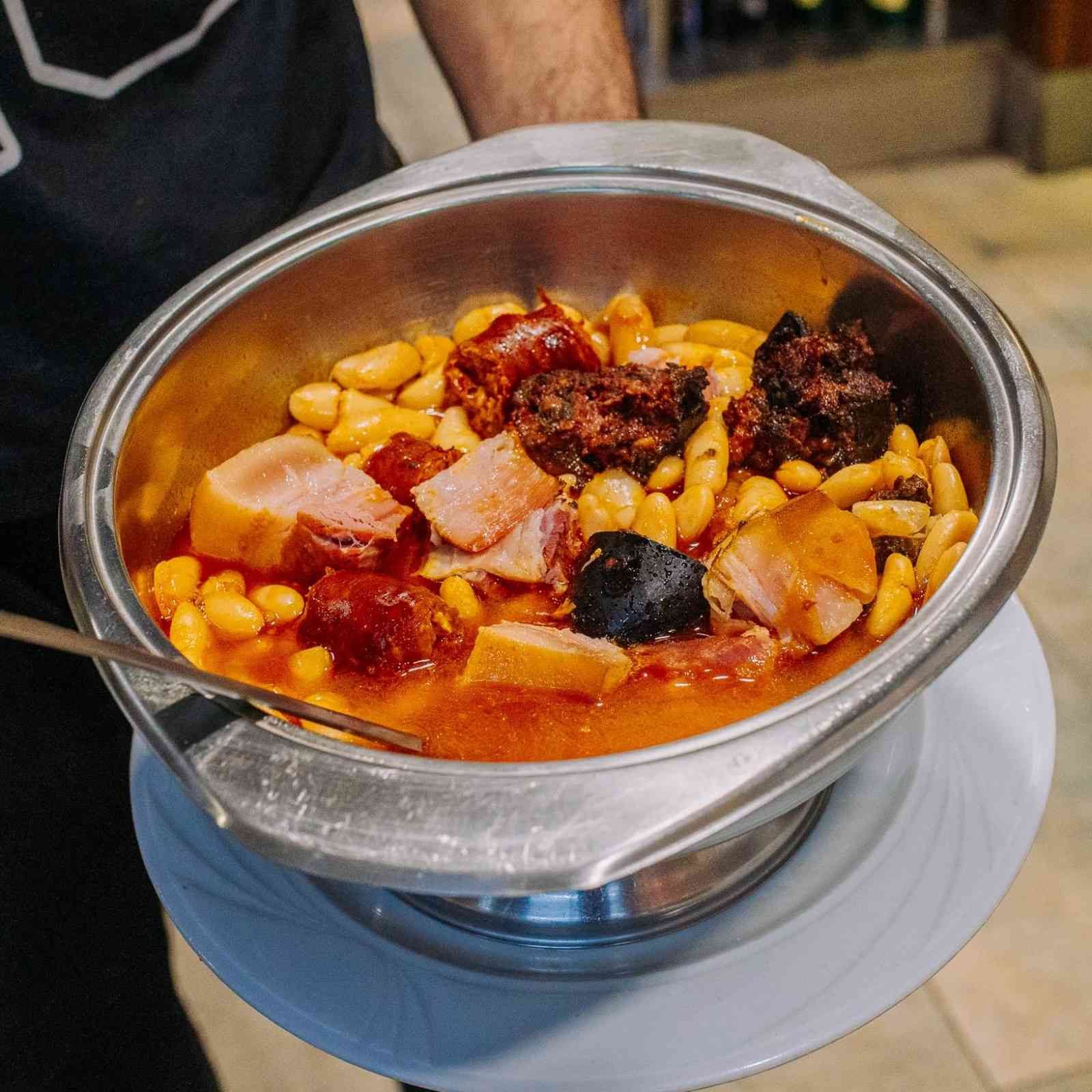 Un tazón de metal con estofado de alubias blancas, salsa roja y trozos de carne sobre una mesa.