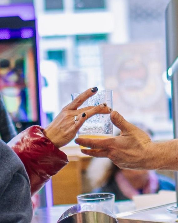 Dos manos intercambian un vaso de plástico transparente en un mostrador, con la colorida fachada de una tienda borrosa al fondo.