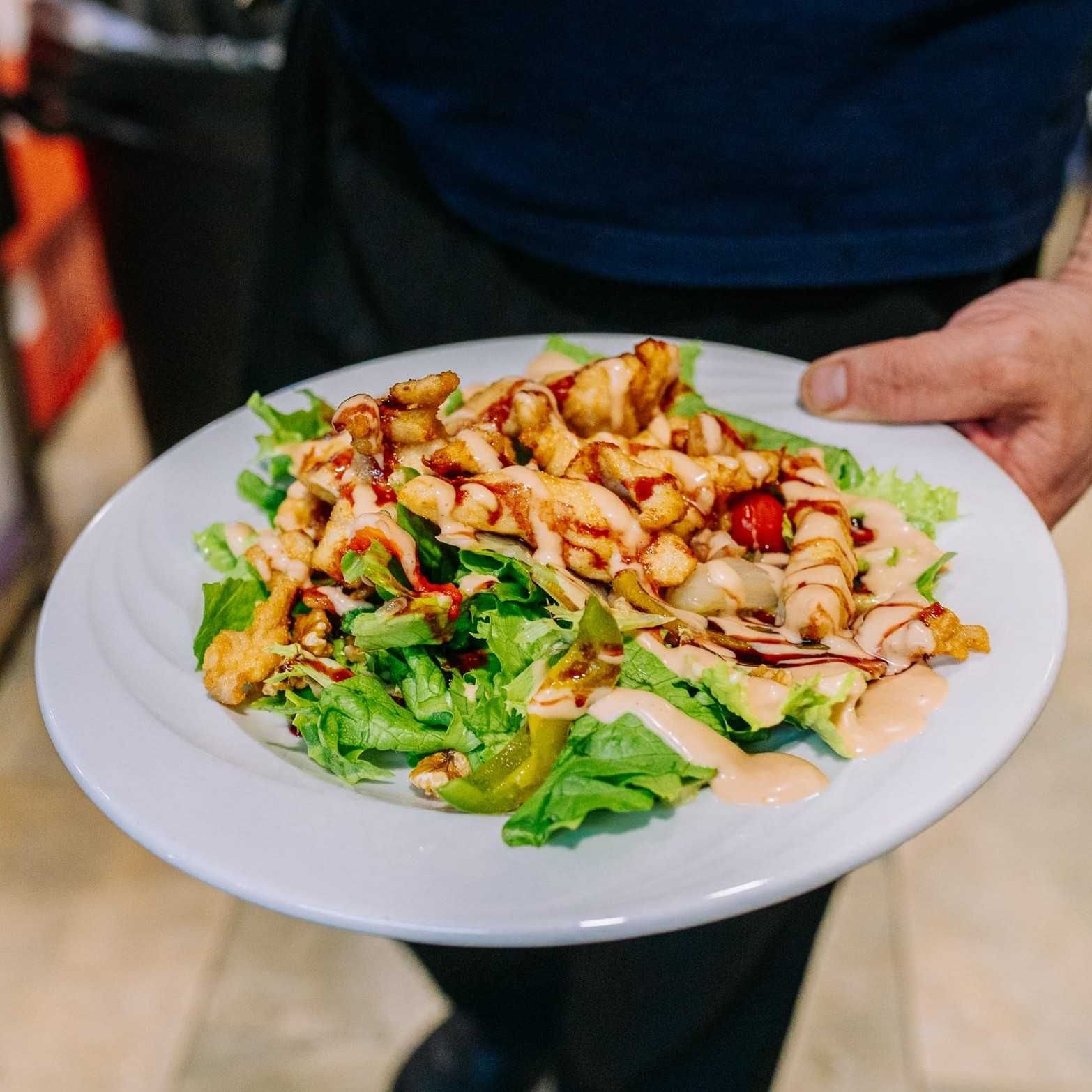 Plato de ensalada con pollo a la parrilla y aderezo, sostenido por una persona con una camisa oscura.