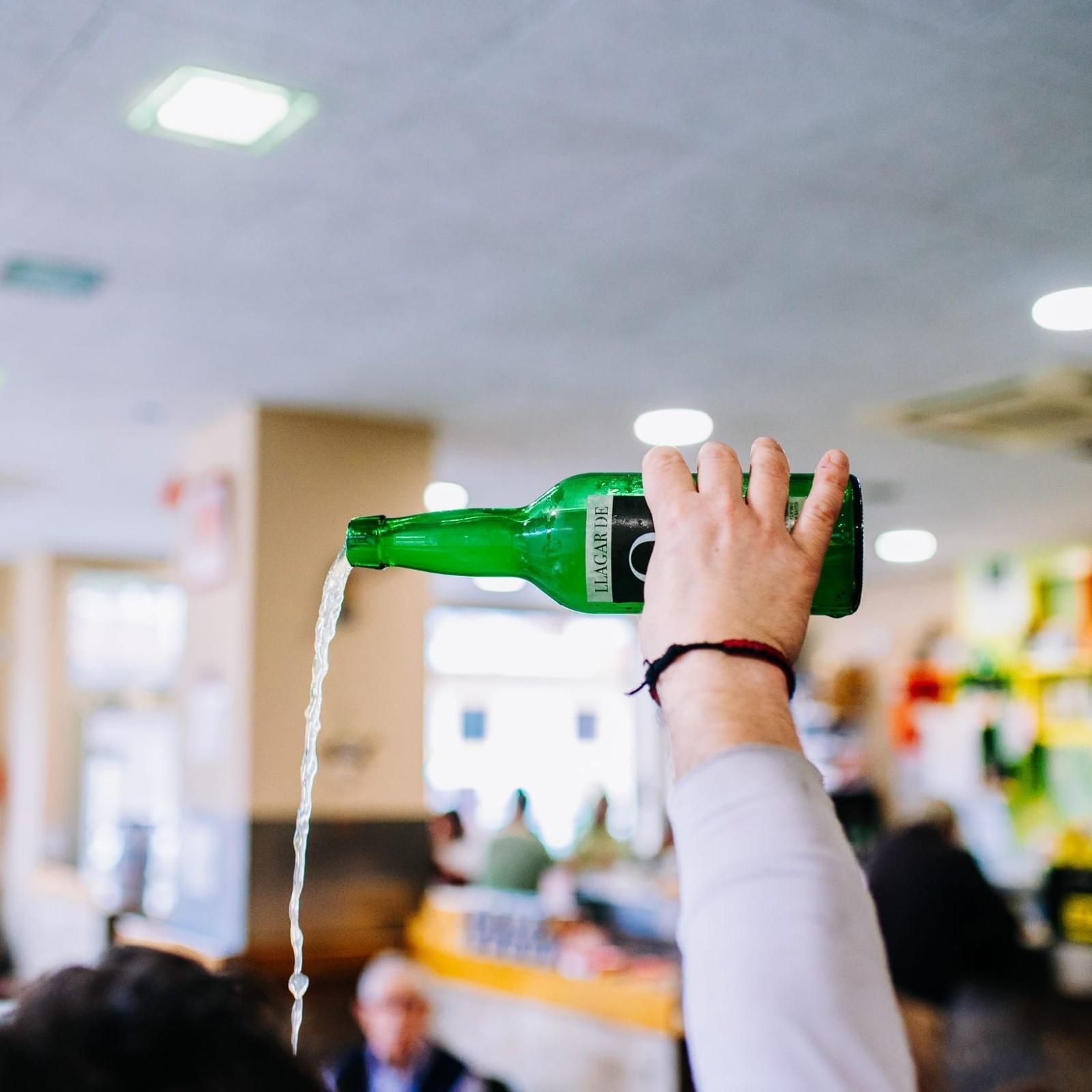Vertiendo líquido a mano desde una botella verde en un entorno interior borroso.