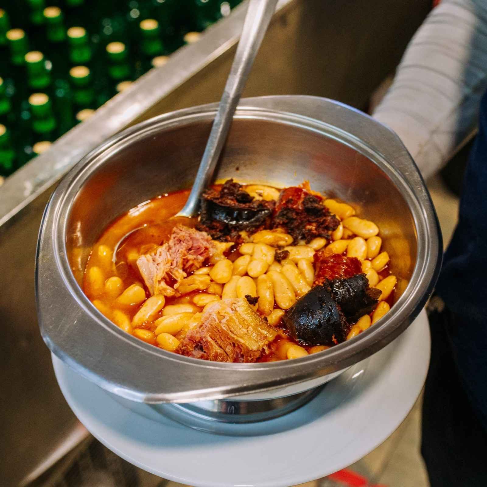 Un tazón de metal con guiso de frijoles, aceite de chile rojo y una cuchara sobre una mesa.