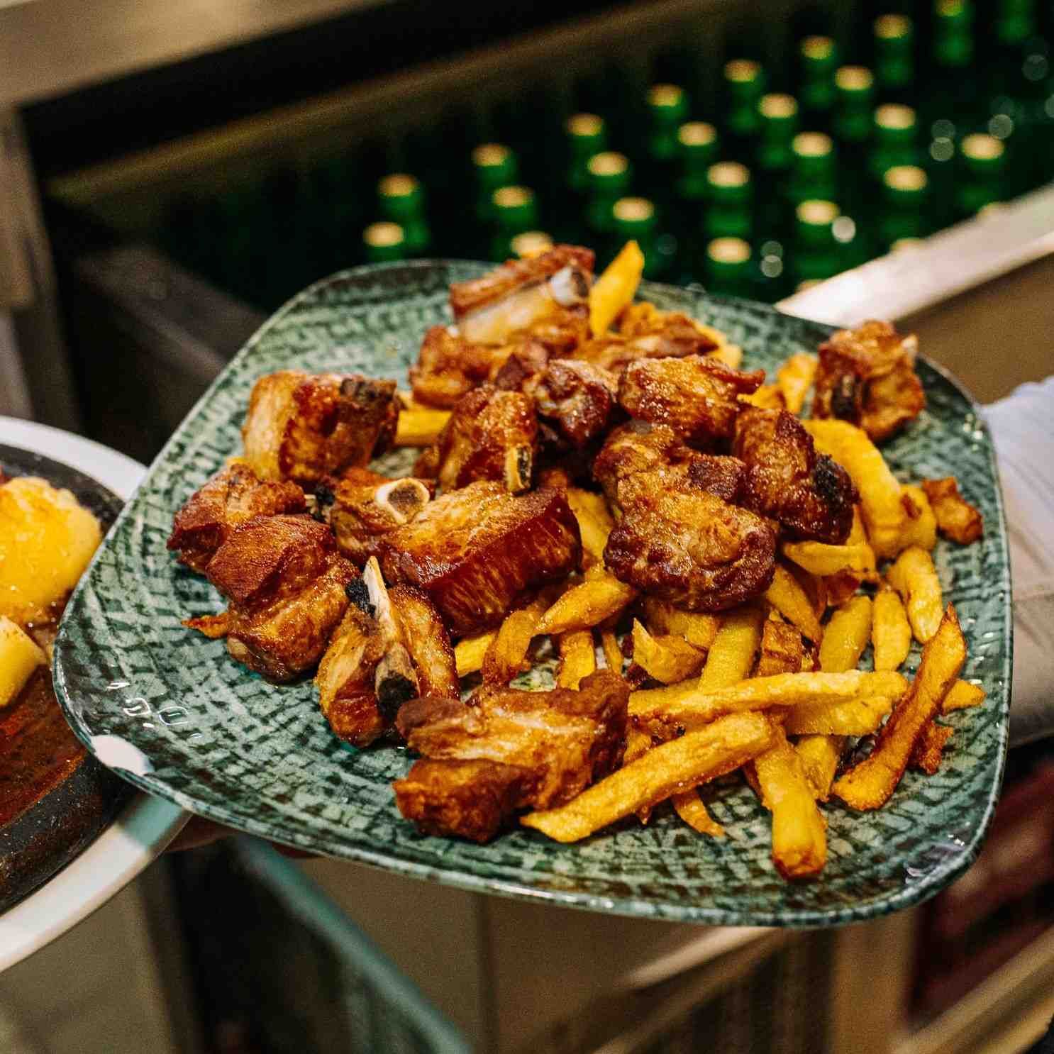 Sartén con patatas fritas y trozos de carne en una bandeja metálica, con botellas verdes borrosas al fondo.