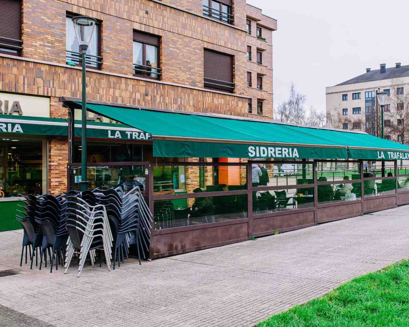 Cafetería al aire libre con toldo verde, sillas negras apiladas y edificios de apartamentos de ladrillo al fondo.