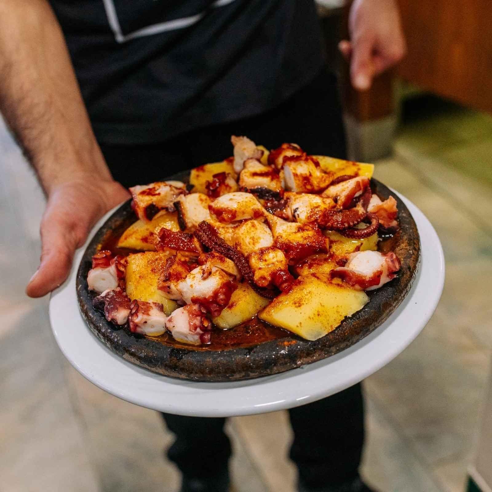 Plato de pulpo a la parrilla y patatas en un plato negro caliente, sostenido con las manos en un restaurante.