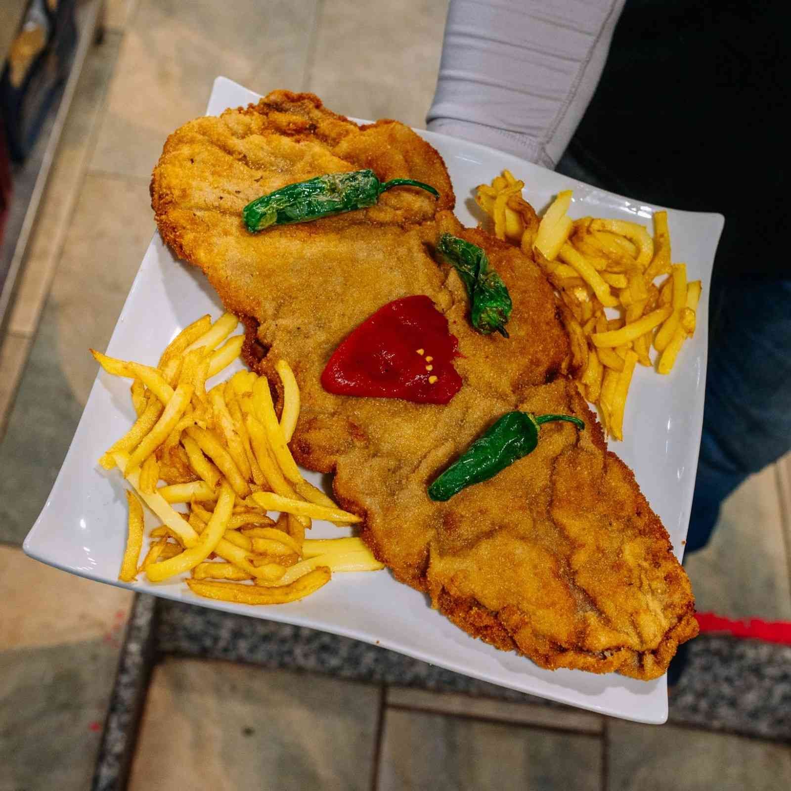 Pescado frito grande con patatas fritas en un plato blanco, cubierto con pimientos verdes y kétchup.