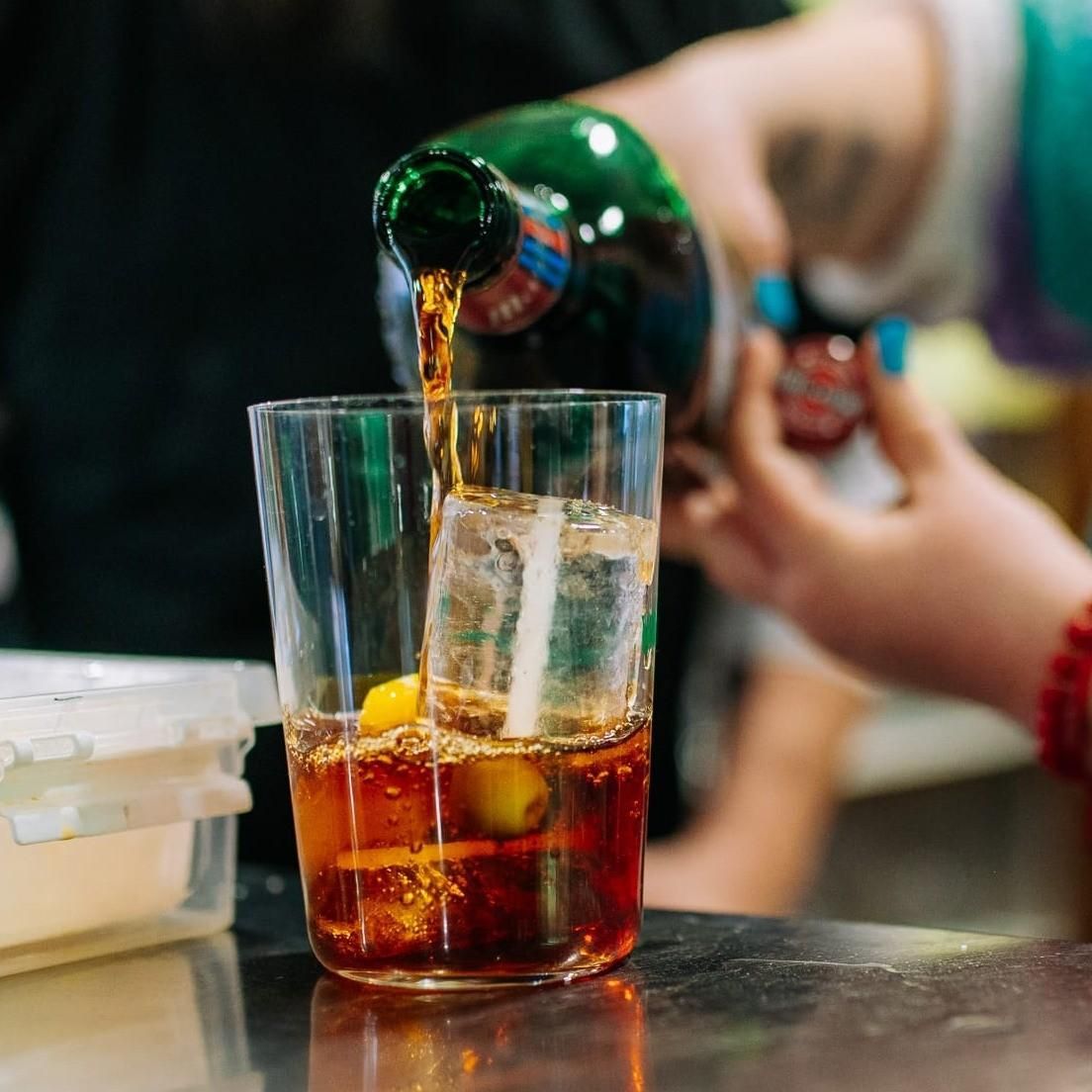 Una mano vierte refresco en un vaso con hielo sobre una mesa, con una persona borrosa al fondo.