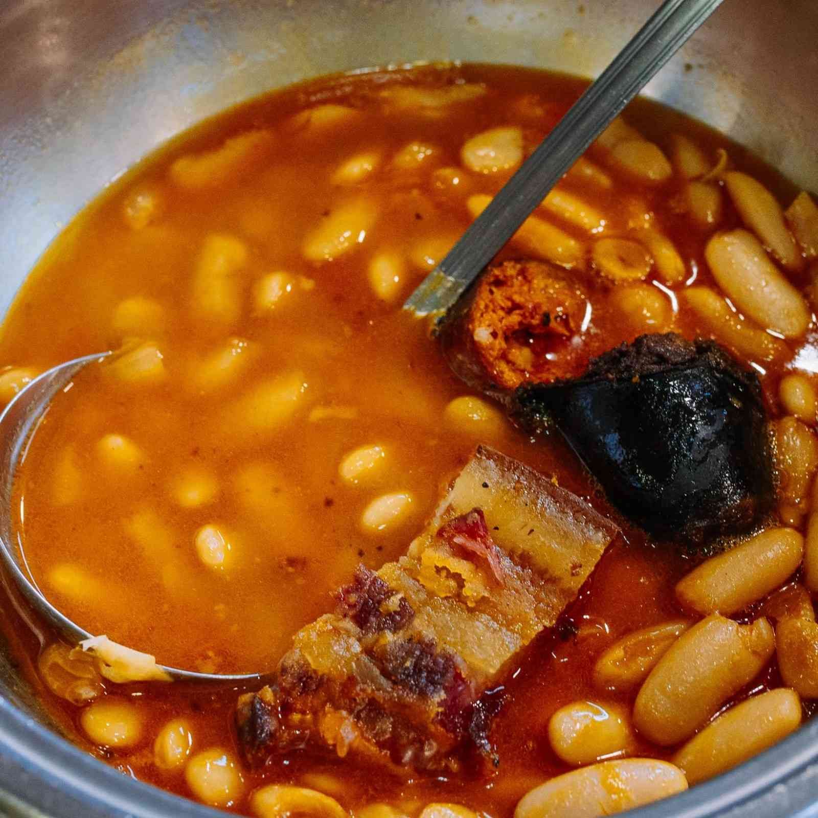 Un tazón de estofado de frijoles con salchicha y trozos chamuscados, caldo de naranja y una cuchara.