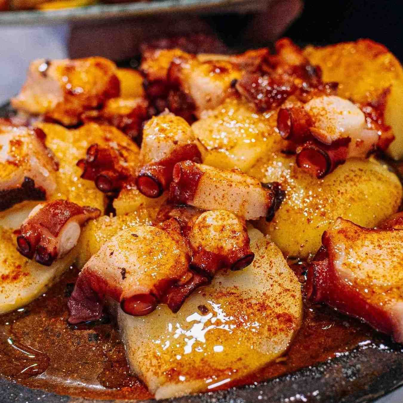 Trozos de pulpo a la parrilla con patatas doradas en un plato oscuro, espolvoreados con pimentón y aceite.