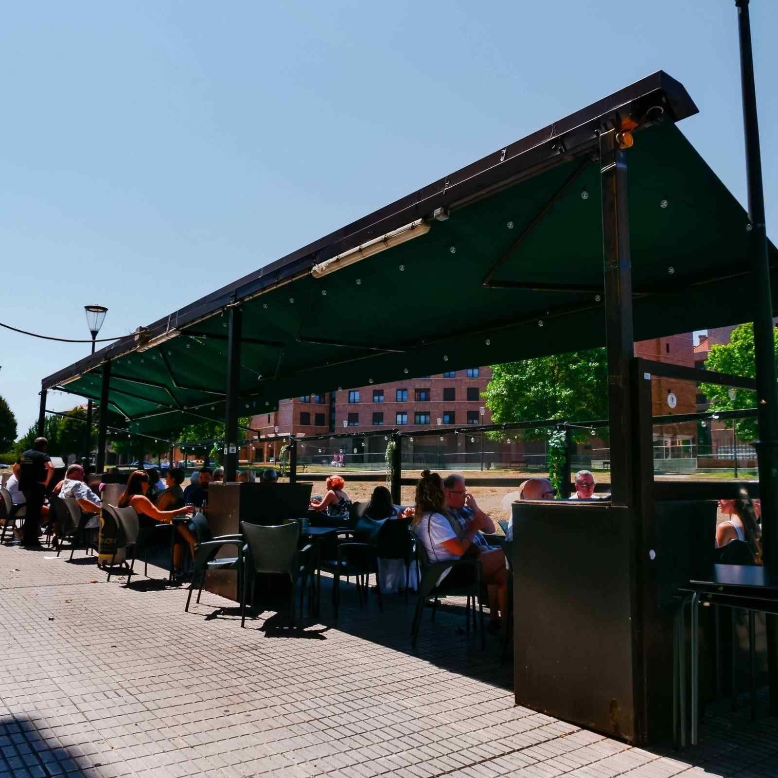 Terraza de una cafetería bajo un toldo verde en una calle soleada, con gente comiendo en las mesas.