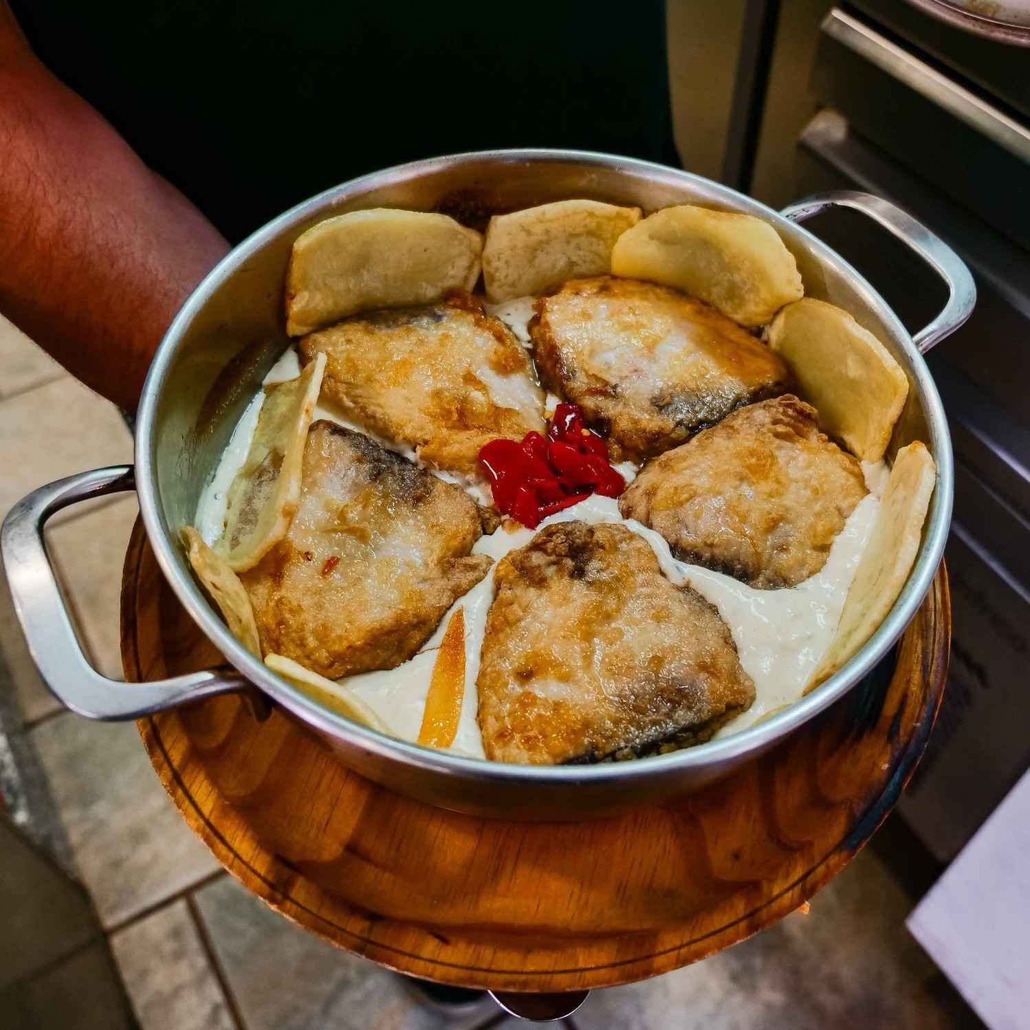 Olla humeante de pescado frito dorado con chiles rojos, rodeada de rebanadas de pan sobre una mesa de madera.
