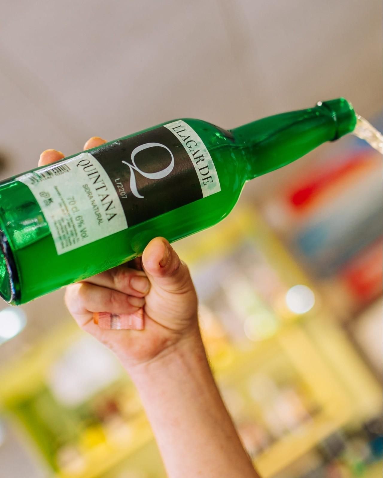 Mano sosteniendo una botella verde de Martini, inclinada contra un fondo de bar borroso.