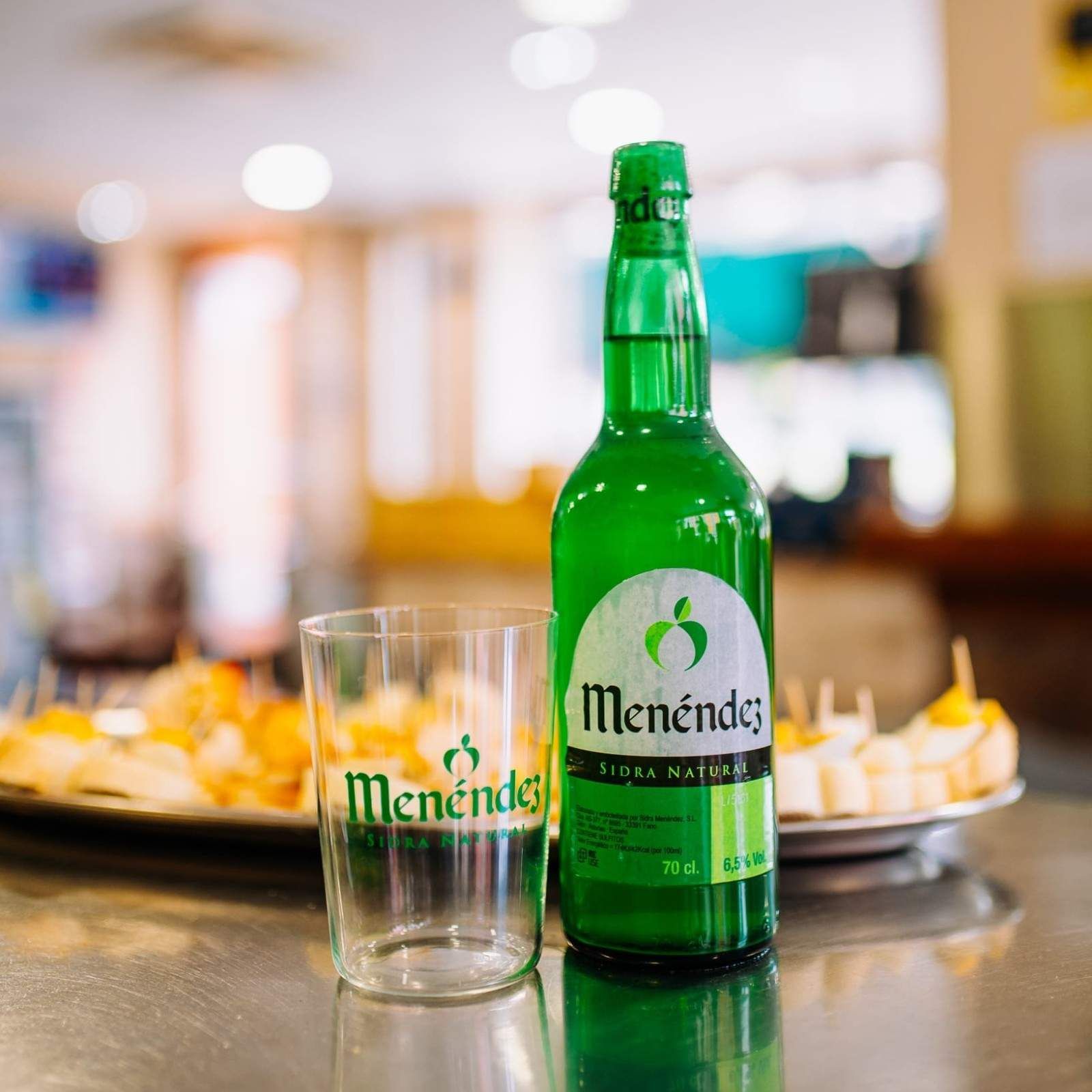 Botella y vaso de Green Menéndez sobre una mesa con bandejas de aperitivos borrosas al fondo.