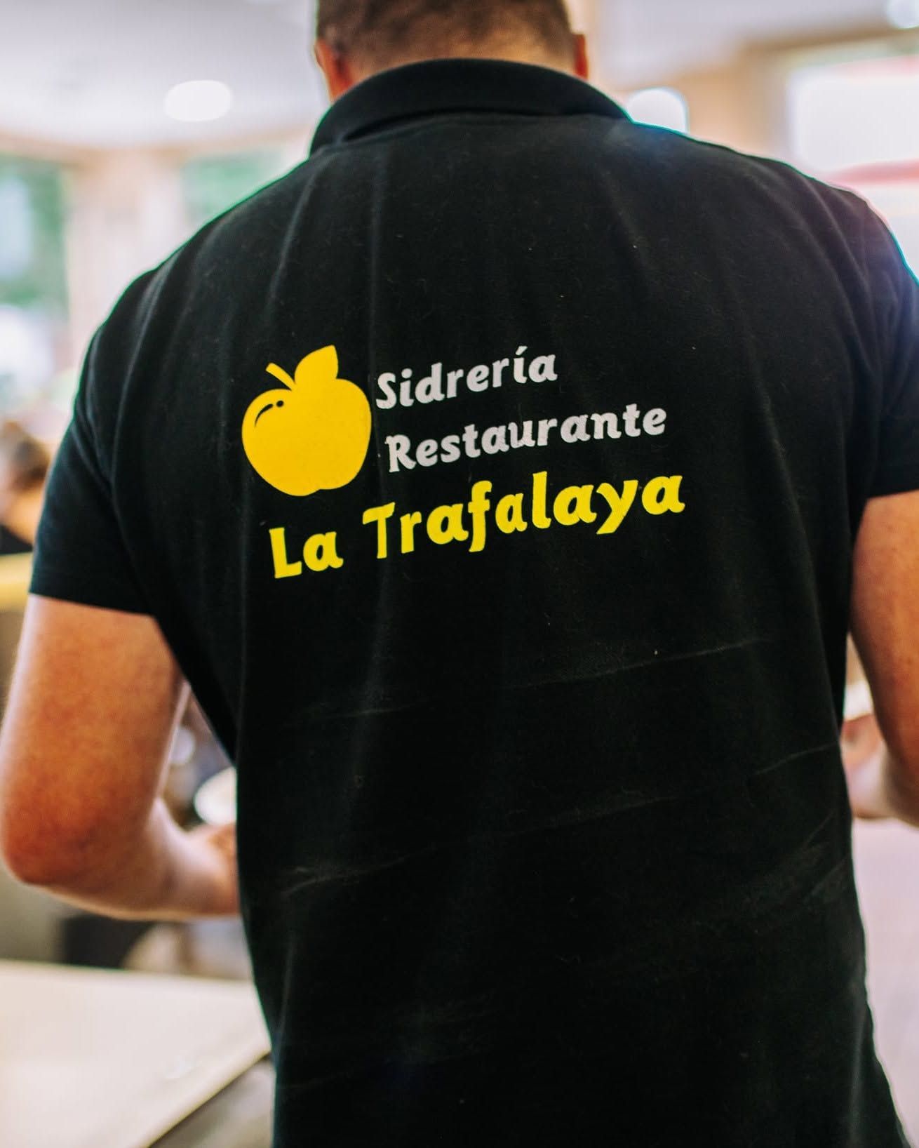 Person wearing a black polo shirt with “Sidrería Restaurante La Trafalaya” logo on the back