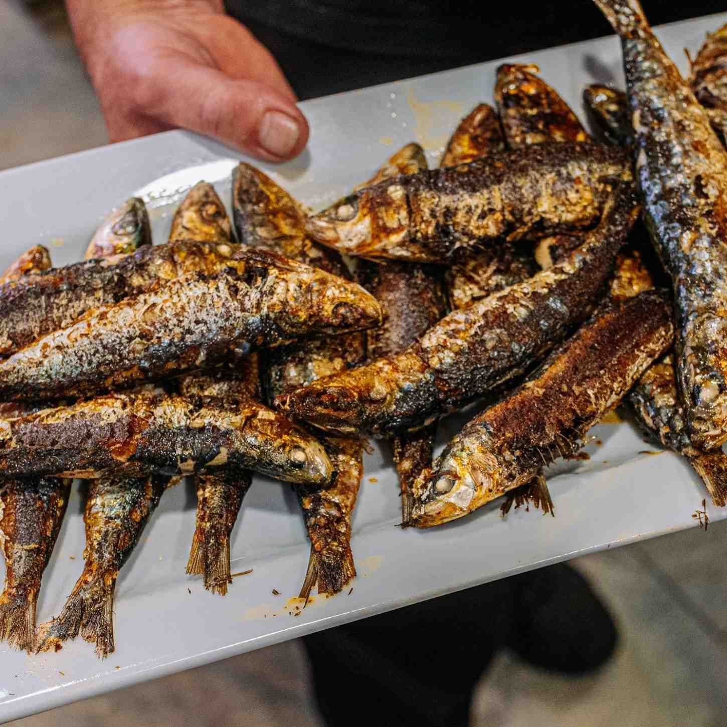 Plato de sardinas a la parrilla con piel crujiente, sostenidas en una mano sobre una bandeja.