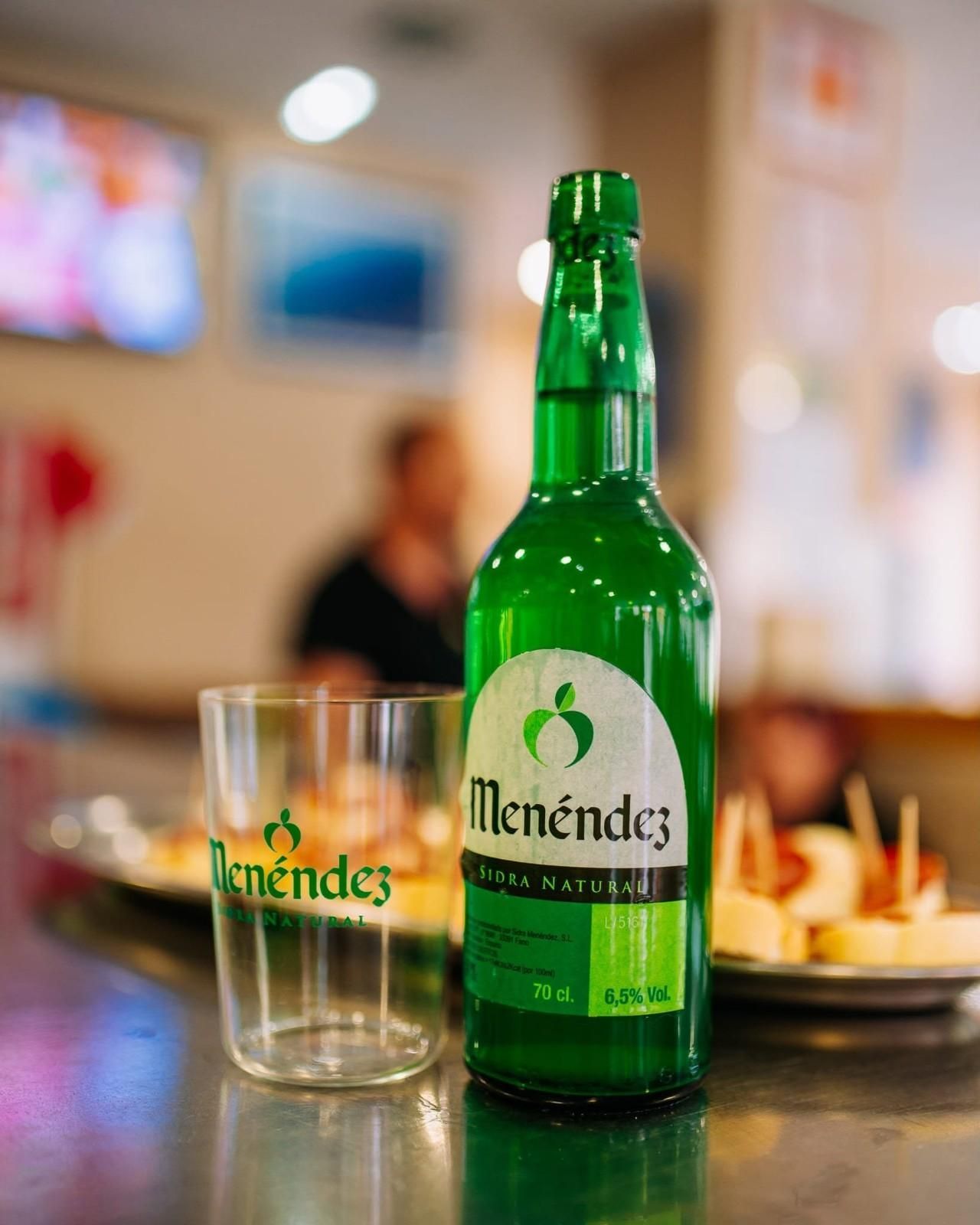 Botella y vaso de Green Menéndez sobre la mesa de un restaurante, con comensales borrosos al fondo.