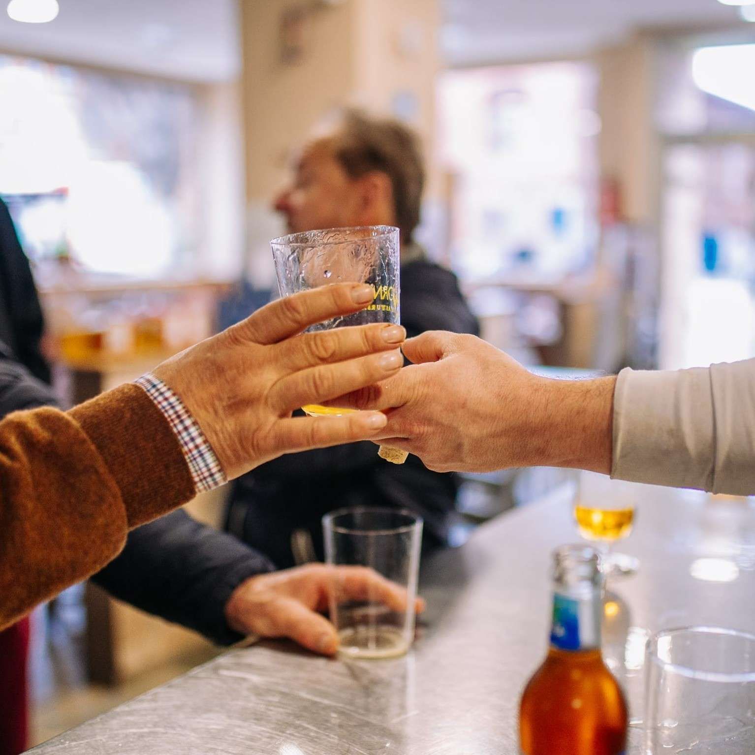 Dos manos brindando con bebidas en la barra de un bar, con una persona borrosa al fondo.