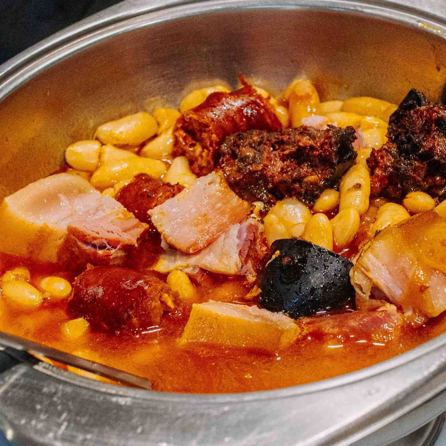 Primer plano de un tazón de metal con estofado de frijoles, trozos de carne y salsa roja.