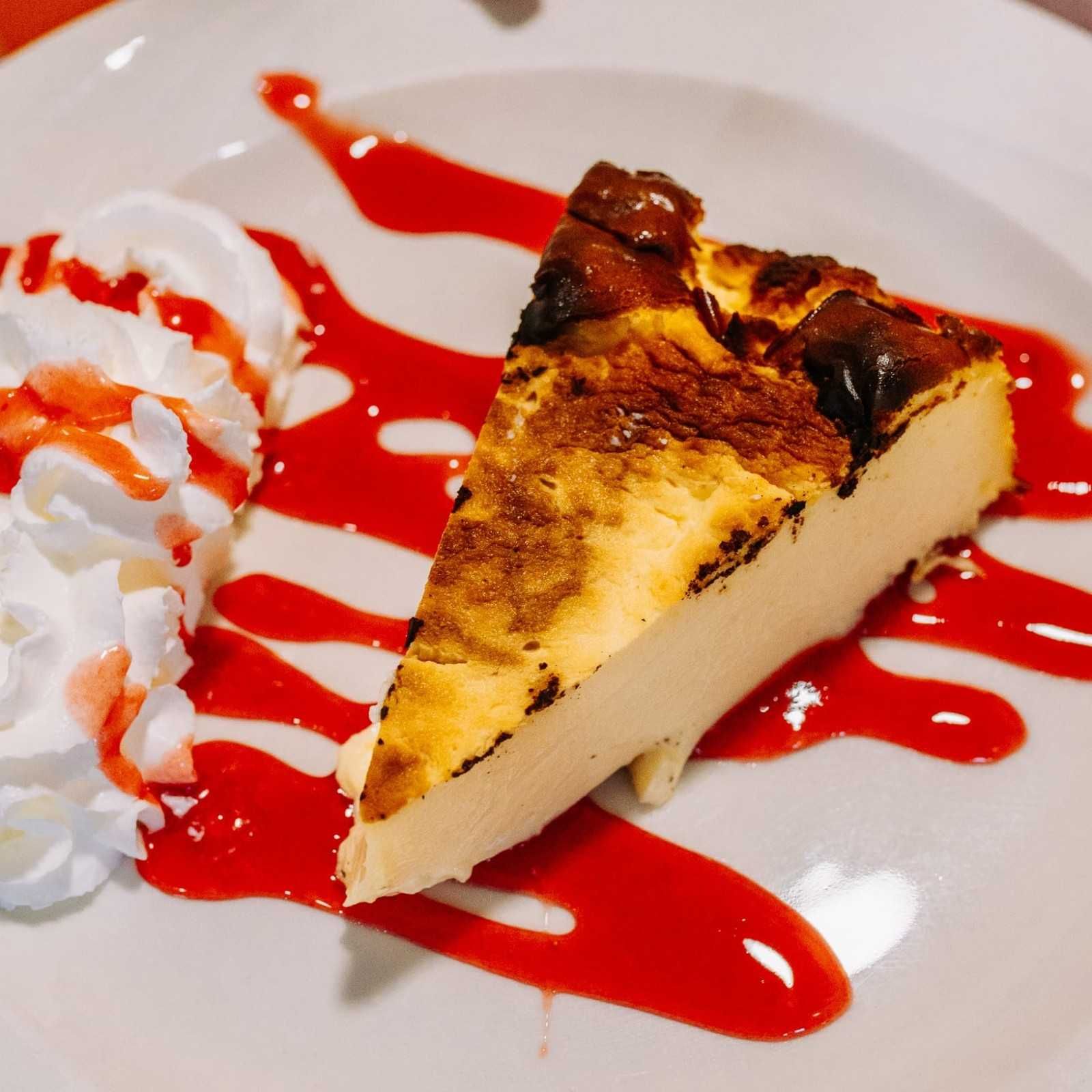 Una porción de tarta de queso bañada en salsa roja y cubierta con crema batida sobre un plato blanco.
