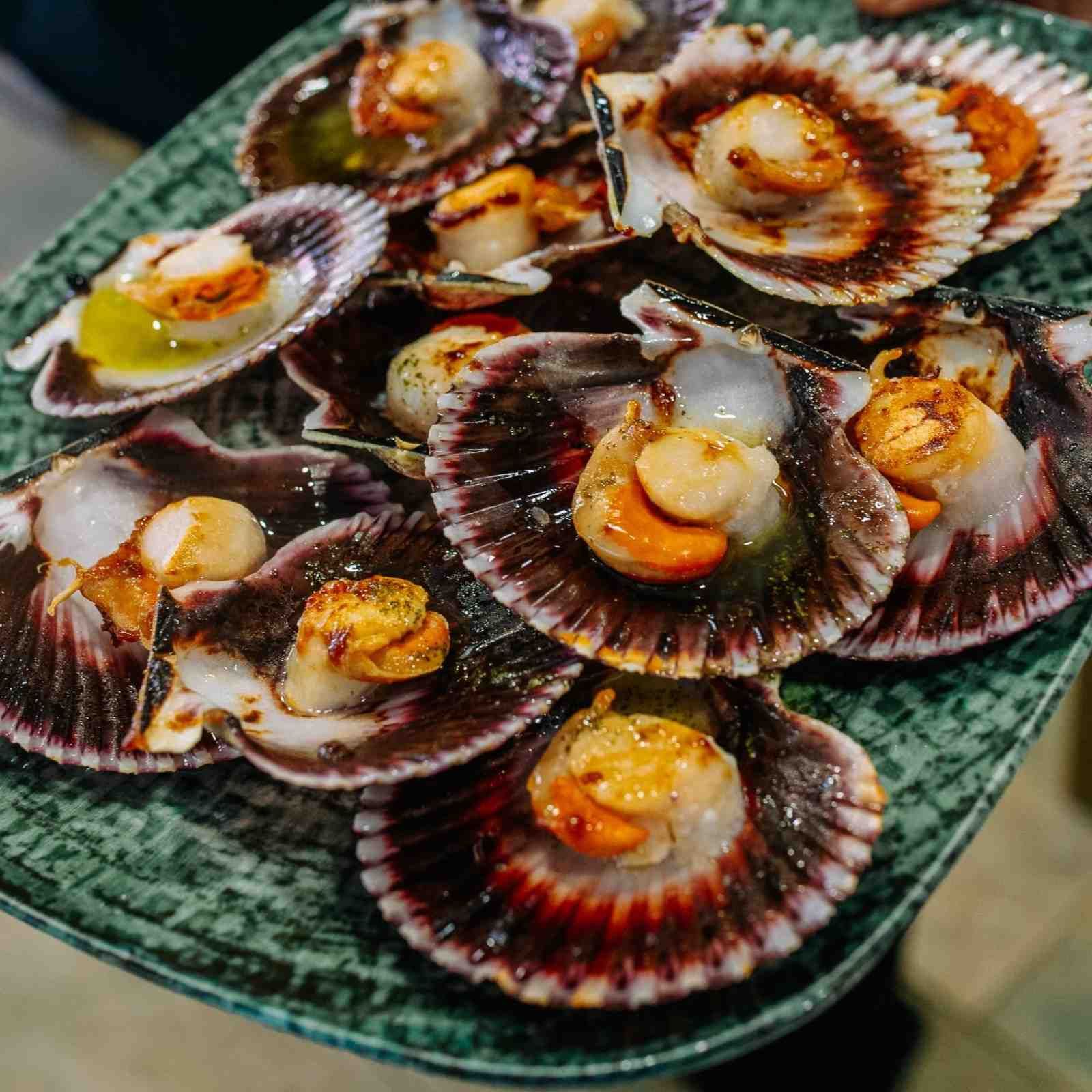 Vieiras a la parrilla servidas en platos de concha, cubiertas con salsa y dispuestas en una fuente verde.