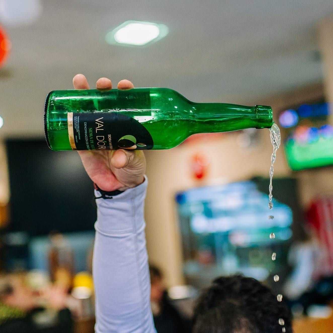 Vertiendo líquido a mano desde una botella verde en el interior de un edificio, con una multitud borrosa al fondo.