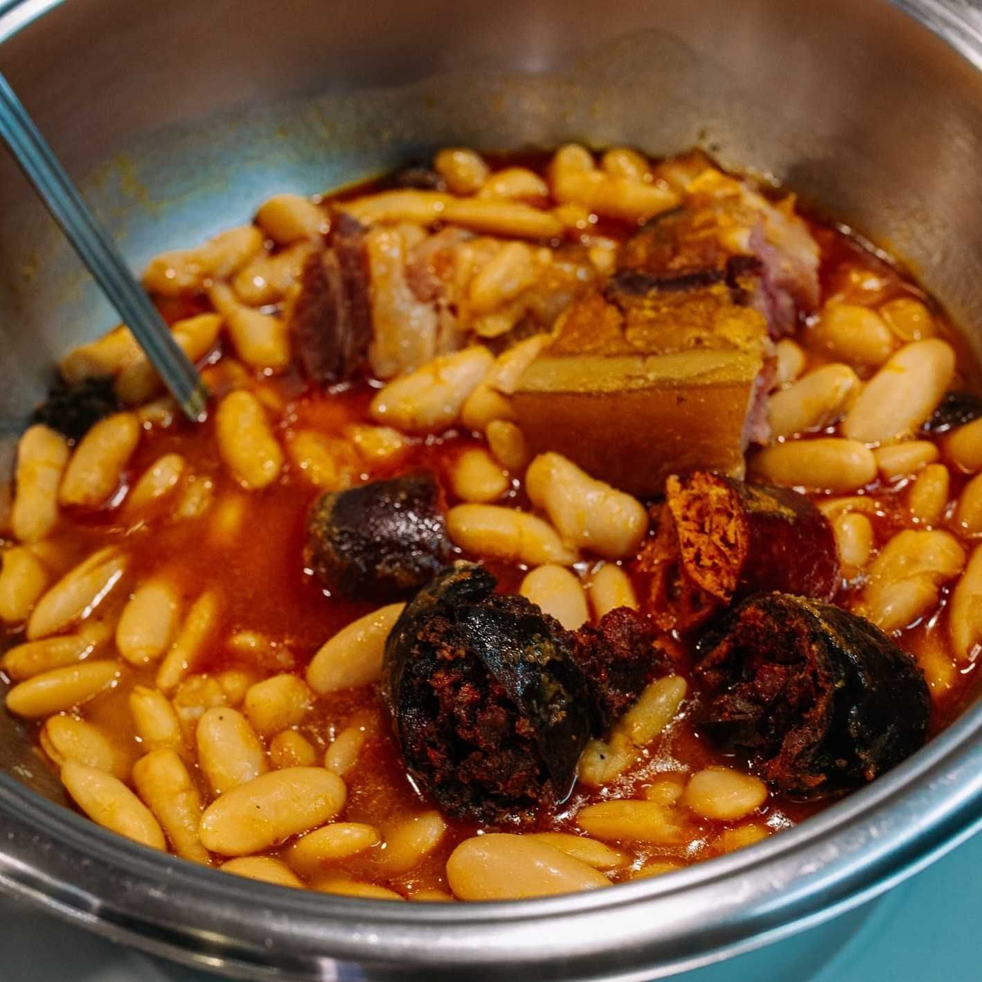Cacerola de metal con frijoles blancos en salsa roja con carne dorada y frutos secos oscuros, removida con una cuchara.