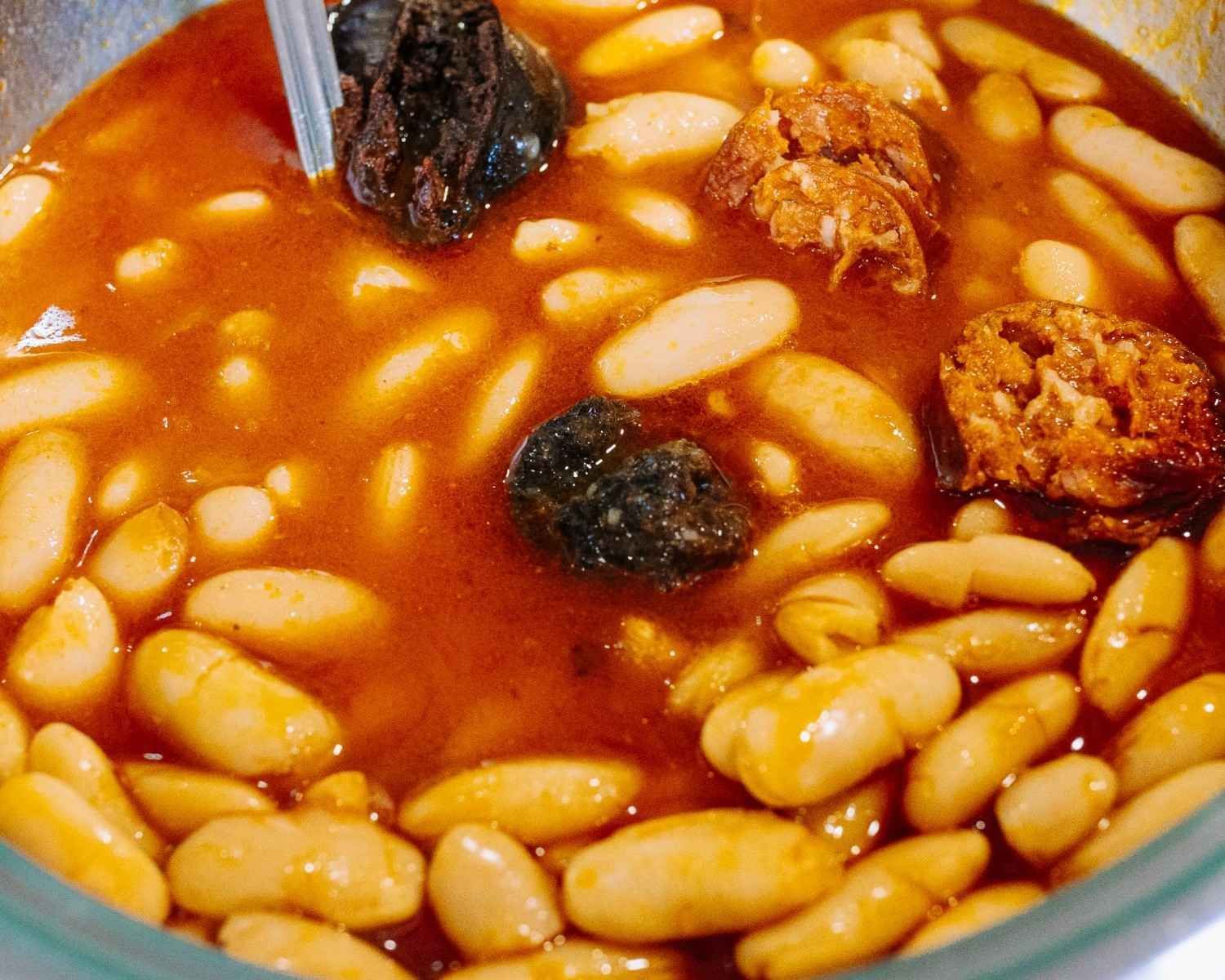 Primer plano de judías blancas en salsa de tomate con trozos de salchicha oscura en un bol.