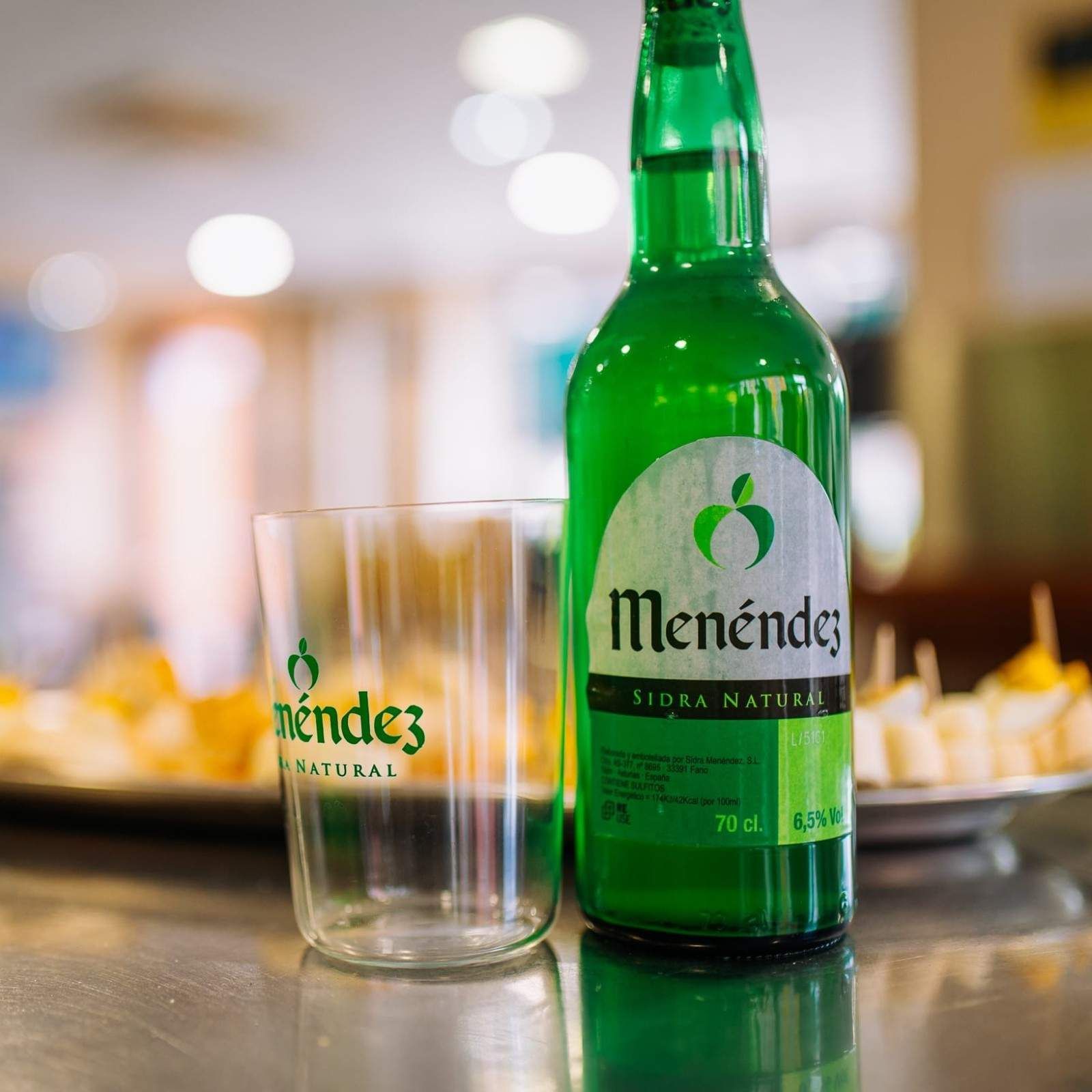 Botella de refresco Green Mendéz y vaso sobre una mesa en un entorno interior borroso.