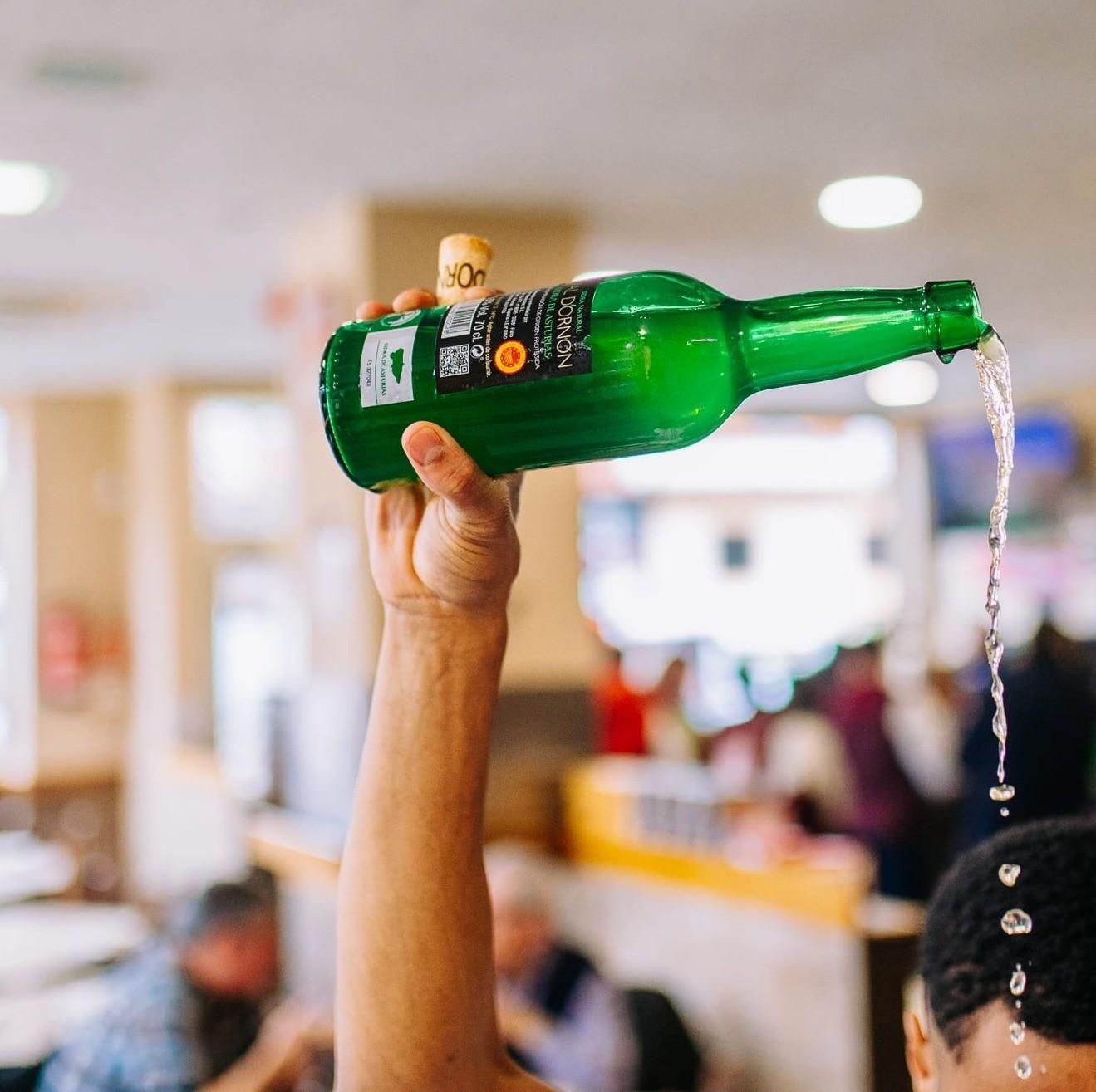 Brazo levantado sosteniendo una botella verde, vertiendo líquido en un entorno interior luminoso.