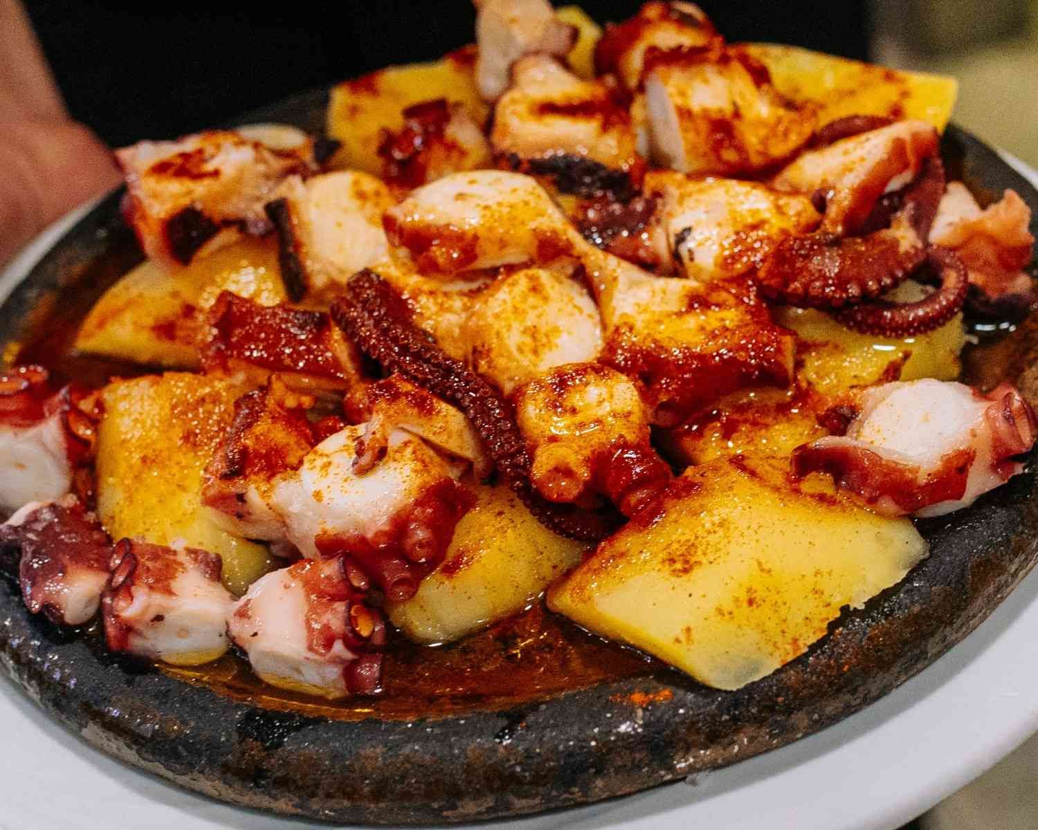 Pulpo a la parrilla con patatas y salsa roja picante en un plato redondo negro.