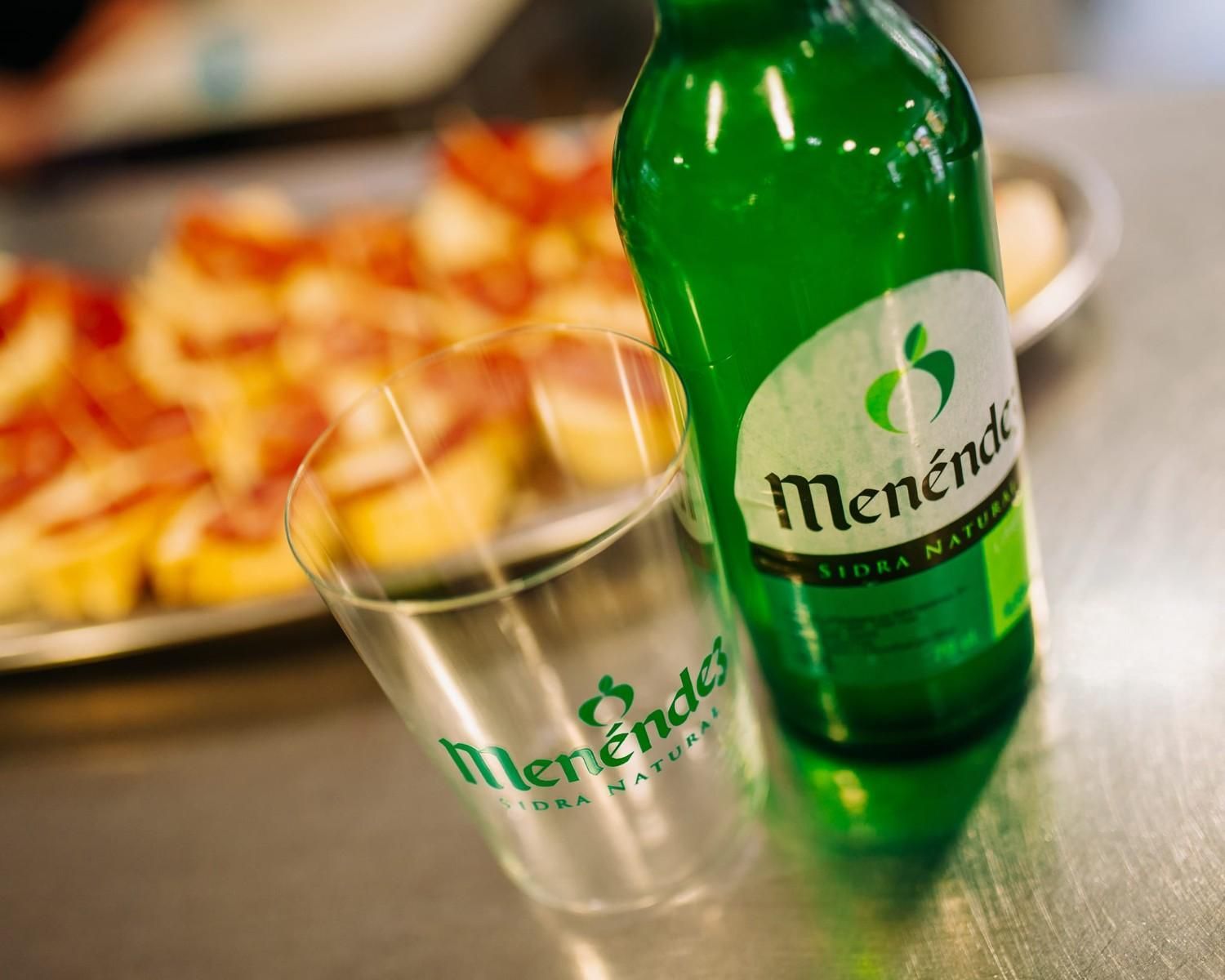Botella y vaso de Mendeta verde sobre una mesa, con un plato de comida borroso al fondo.