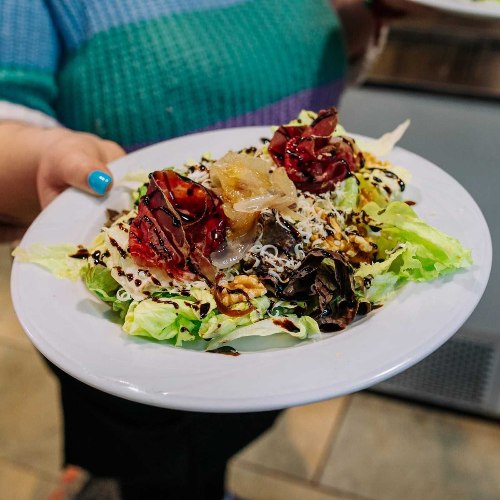 Plato de ensalada con pimientos rojos asados, verduras y queso rallado sobre una mesa, sostenido por una persona con esmalte de uñas azul.