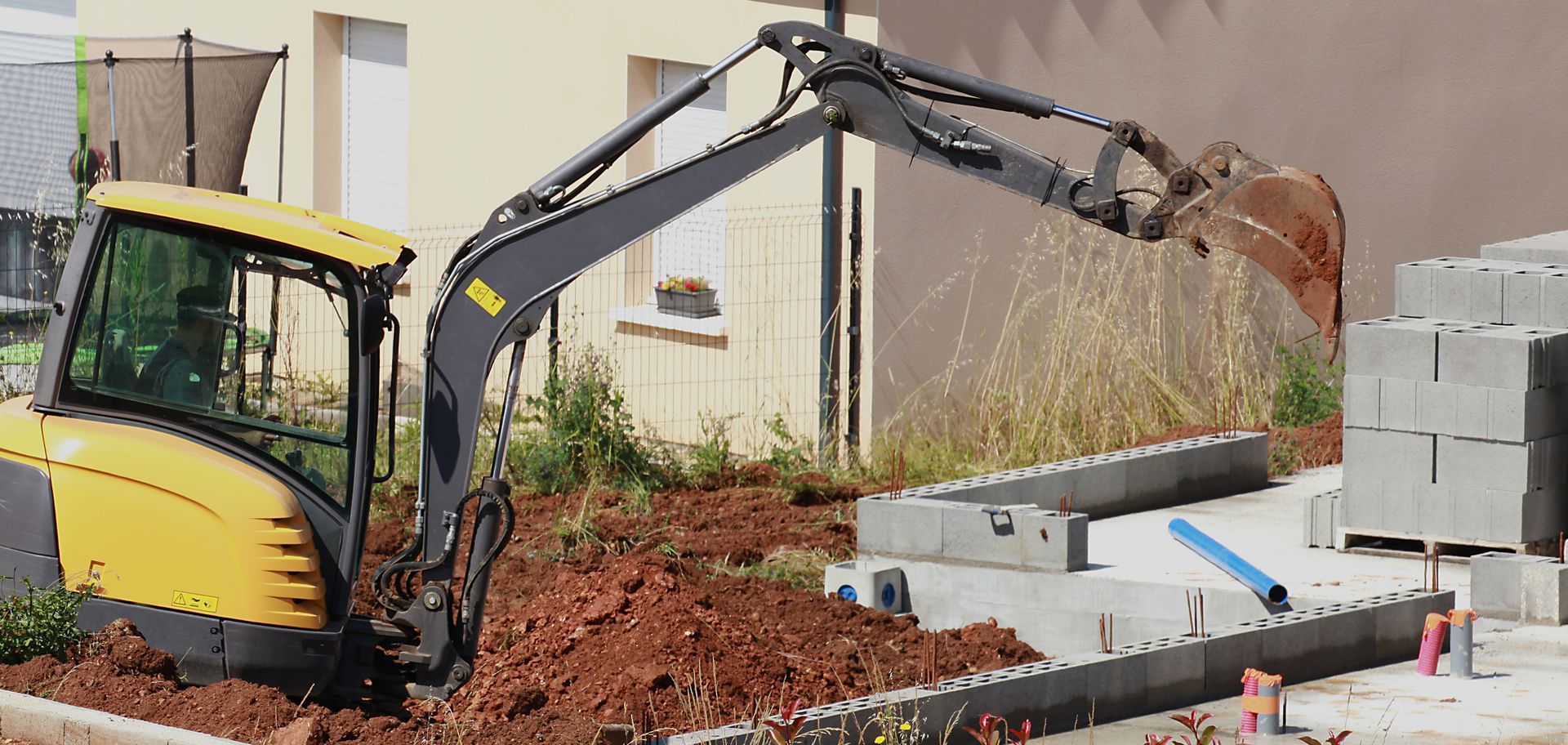 Bras d'excavatrice jaune chargeant de la terre à côté de parpaings empilés sur un chantier de construction.