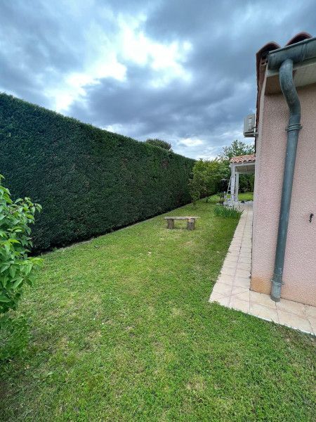 Pelouse arrière bordée d'une haute haie et d'un mur de maison sous un ciel nuageux.