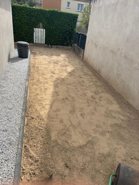 Cour arrière étroite en terre battue entre les murs de la maison, bordée de gravier, avec une haie et un portail au fond.